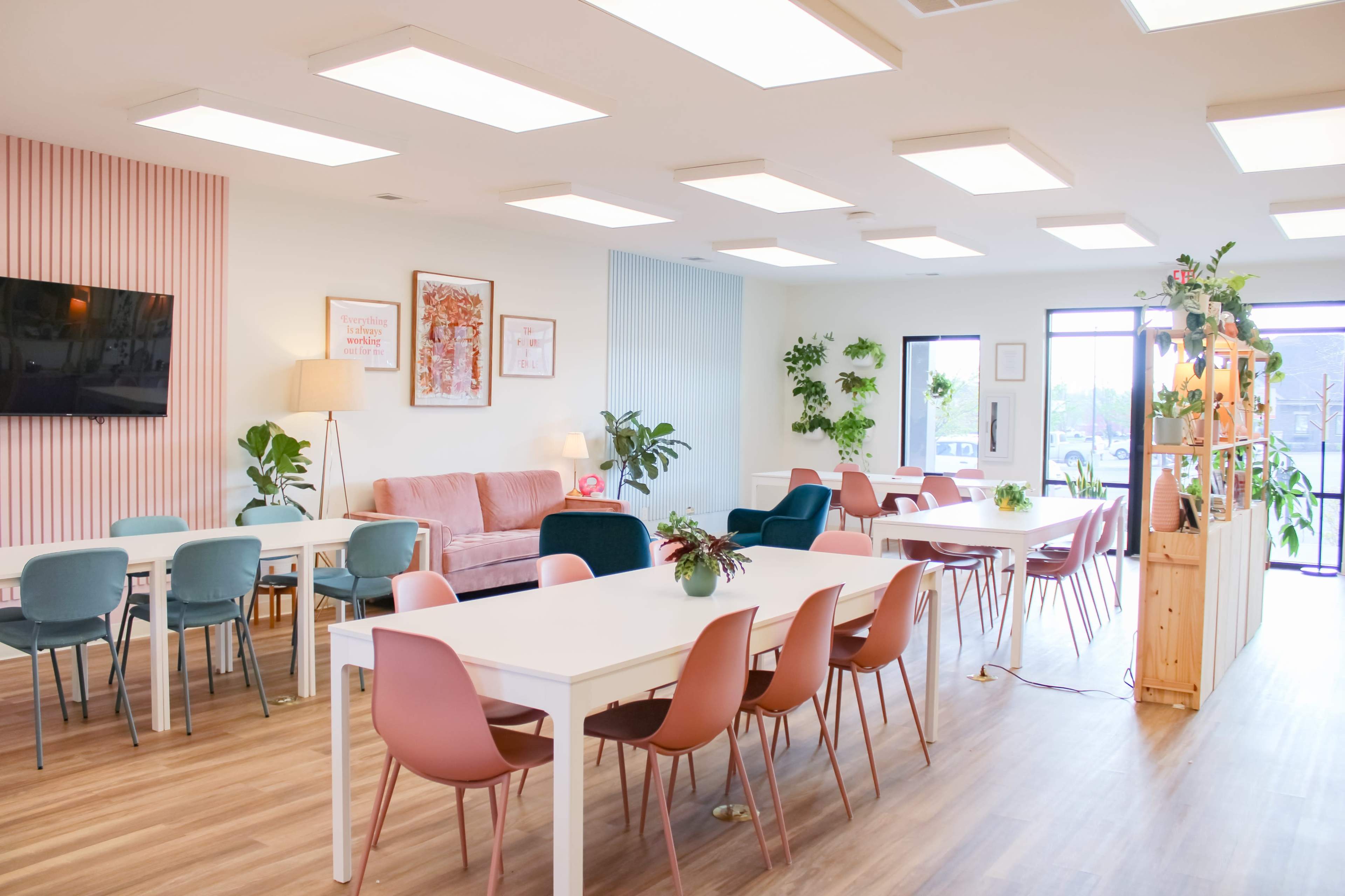 The image shows a bright and modern co-working space featuring white tables, colorful chairs, and large windows with plants decorating the interior.