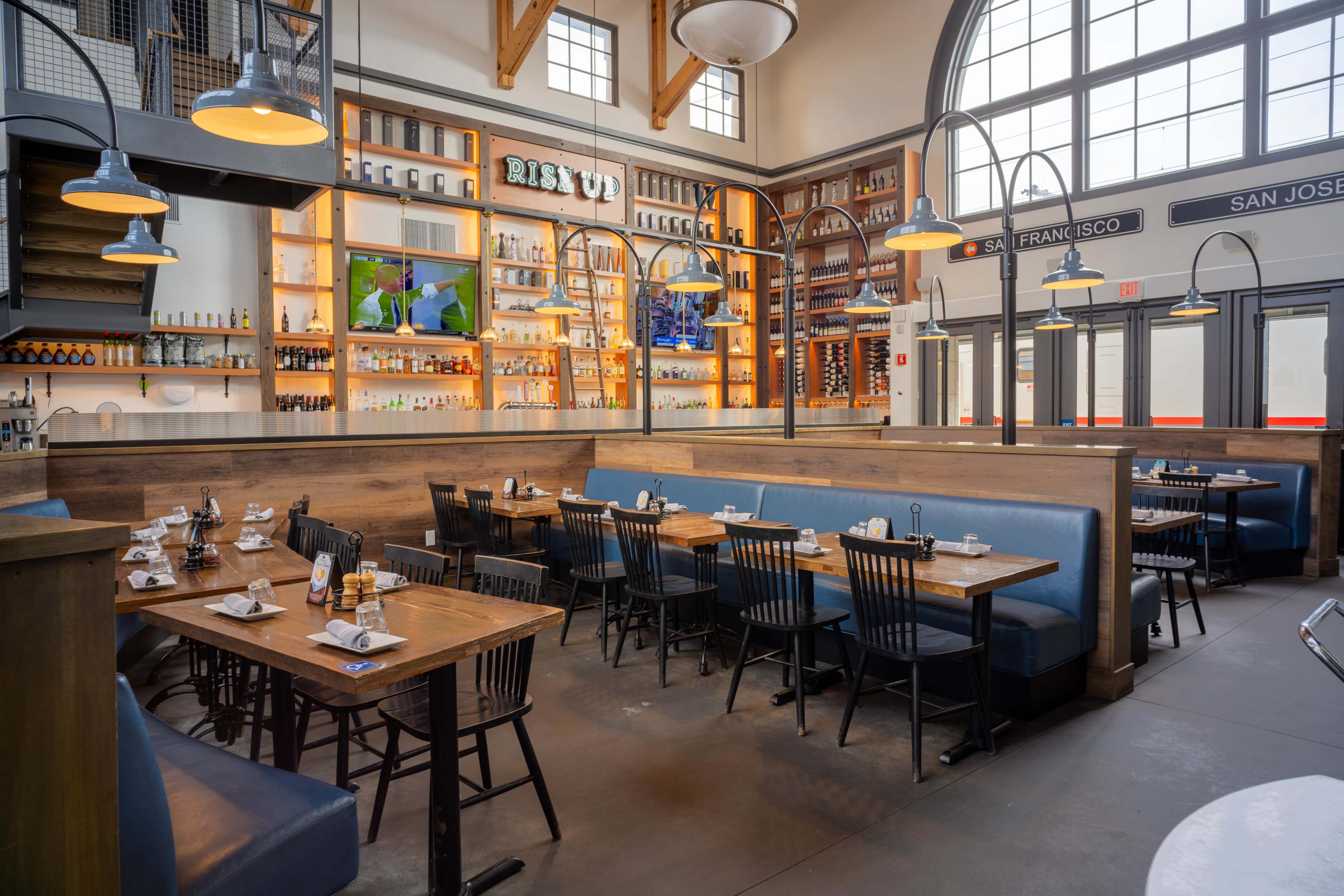 The image shows a spacious restaurant interior featuring wooden tables, black chairs, and a well-stocked bar illuminated by warm lighting.