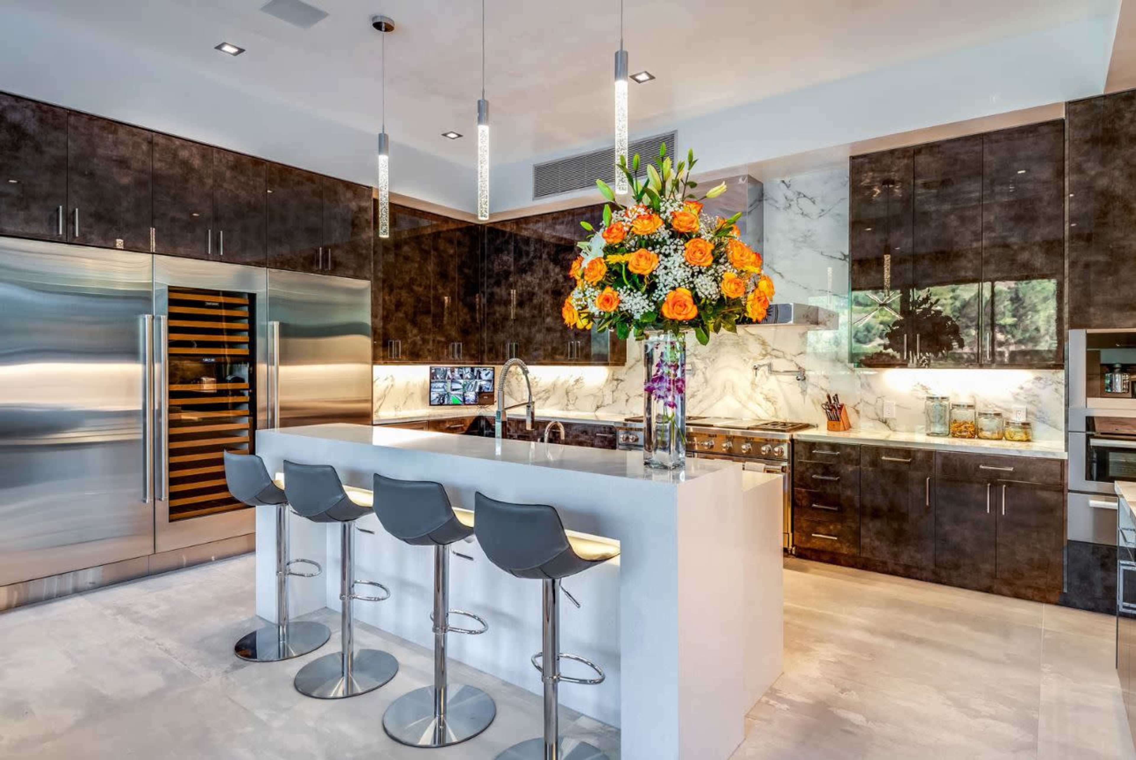 The image shows a modern kitchen featuring dark cabinetry, a large white island with bar seating, stainless steel appliances, and a floral centerpiece.