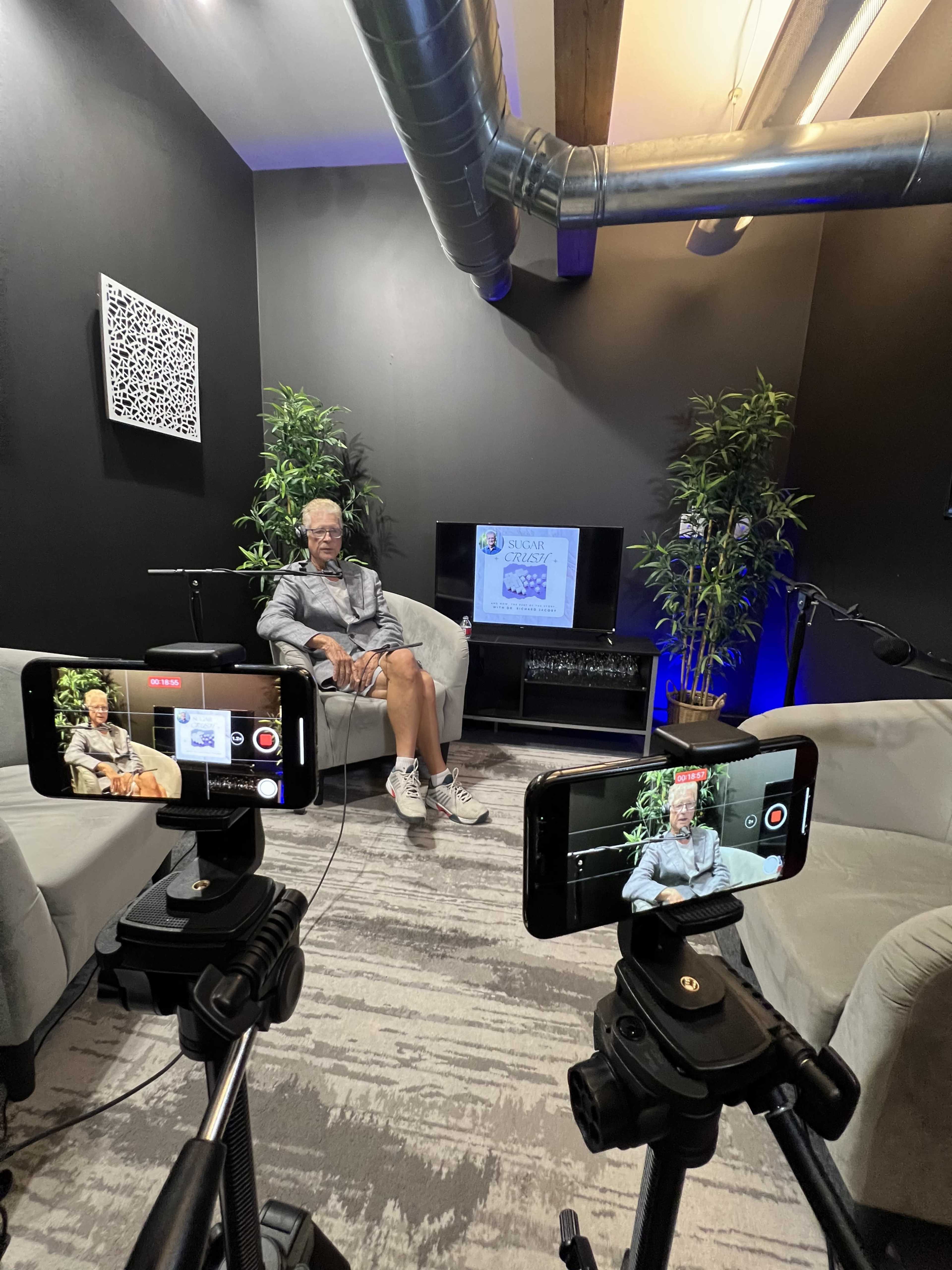 A man sits on a couch in a dimly lit room with plants and a screen displaying content, while cameras on tripods are positioned in front of him.
