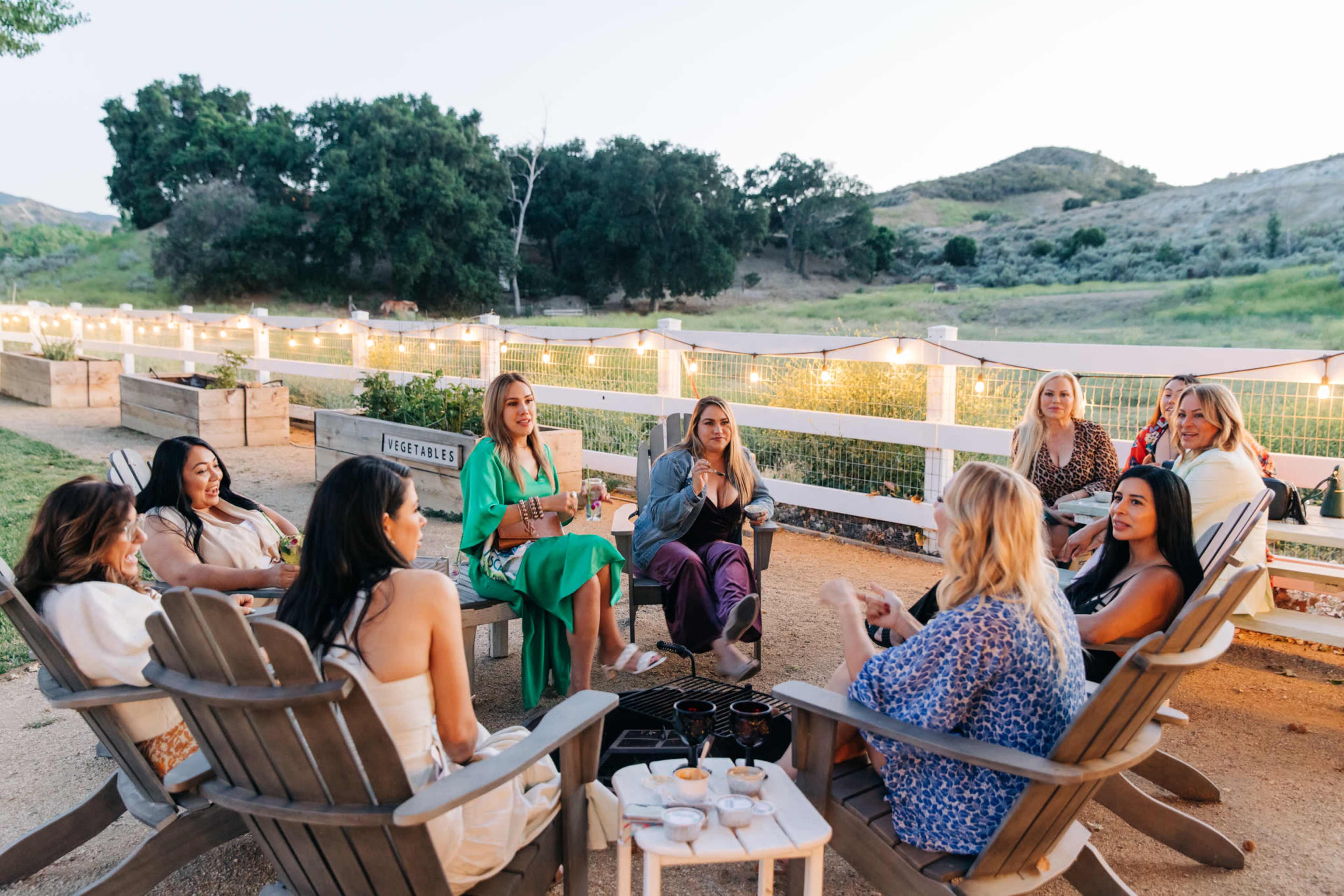 A group of women sits around a fire pit in a relaxed outdoor setting with string lights illuminating the area.