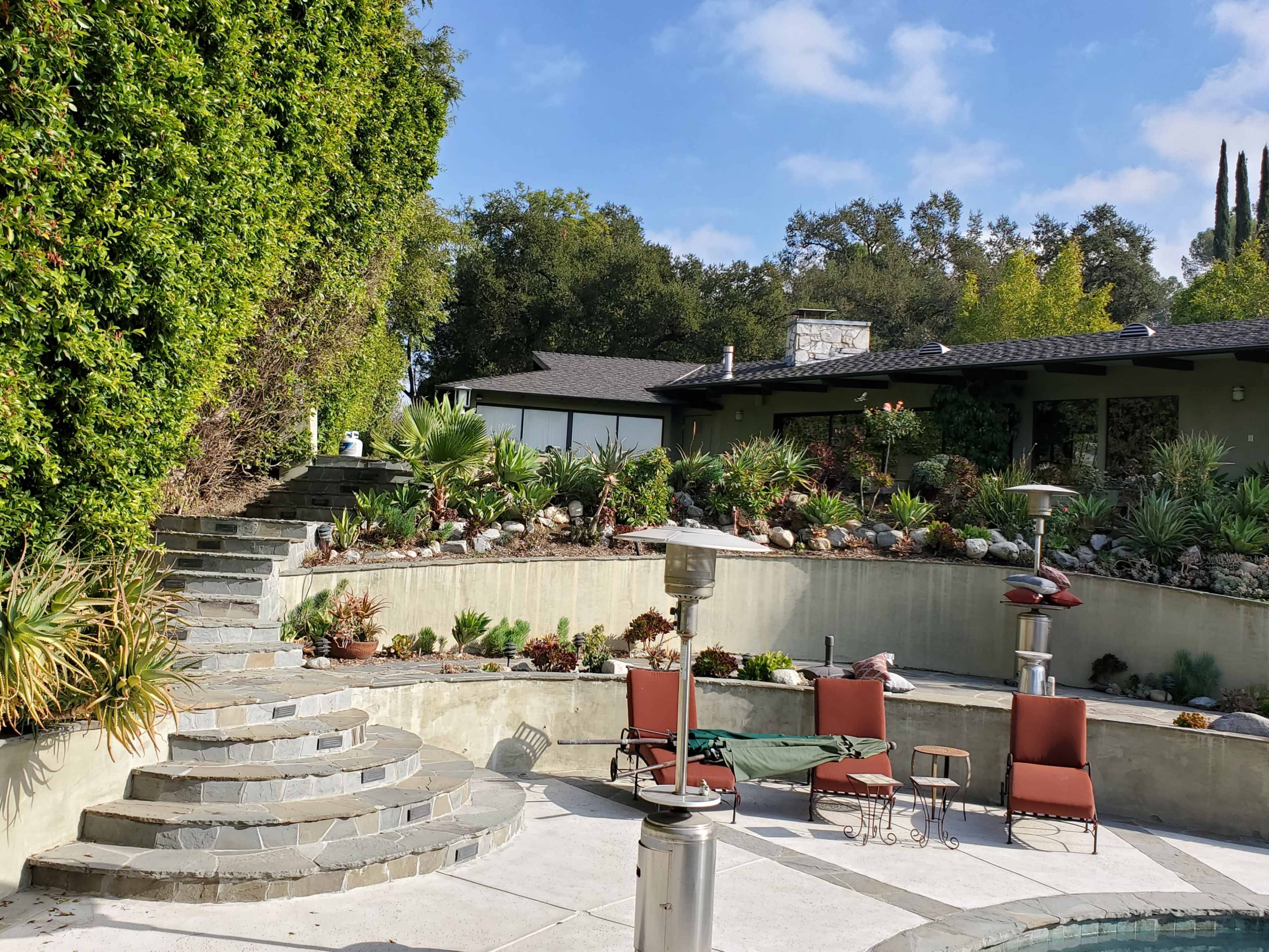 A landscaped backyard with a stone staircase leading to a pool area, surrounded by greenery and patio furniture.