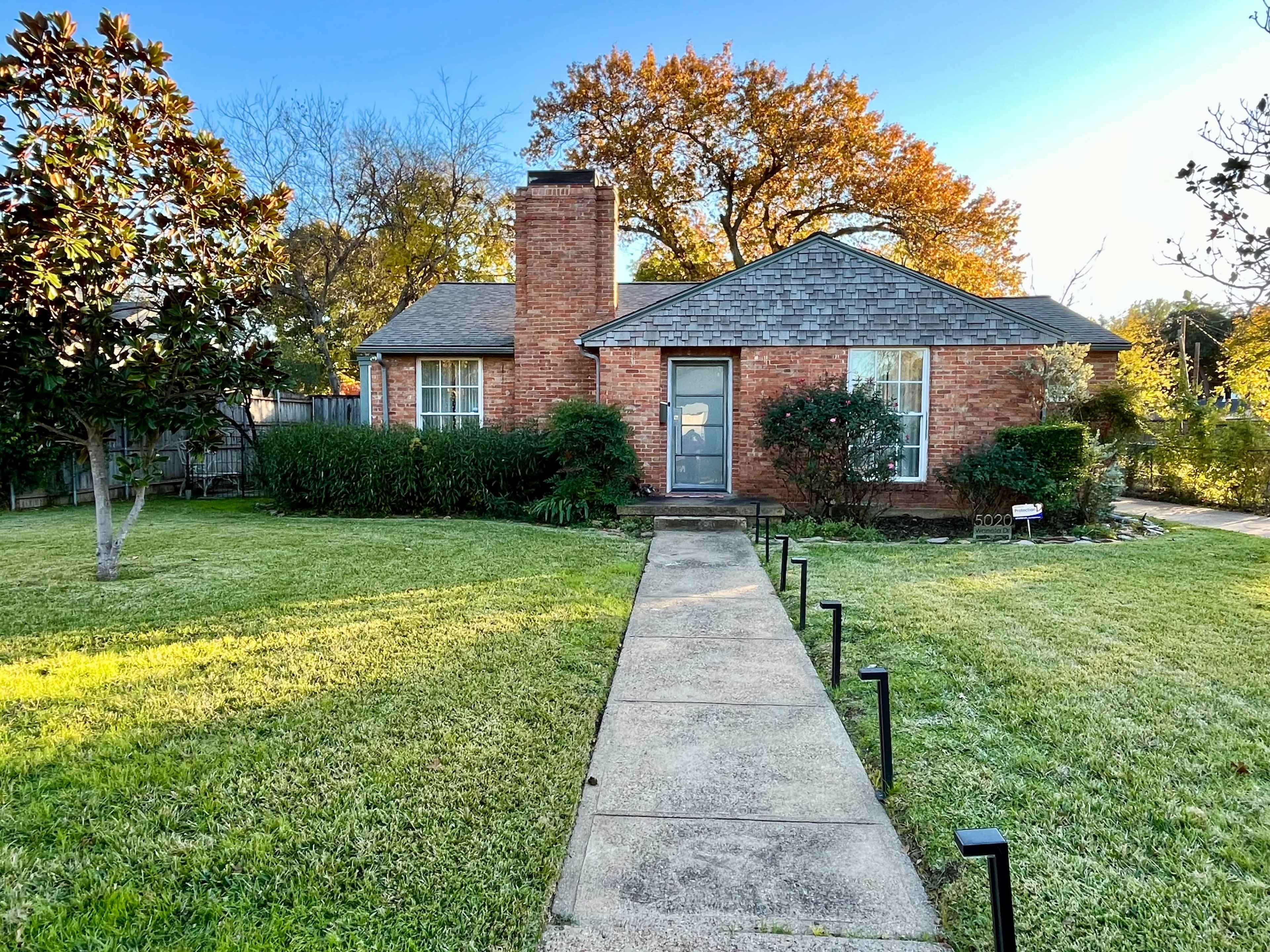 A brick house with a chimney and a front path is surrounded by a well-maintained lawn and shrubs.