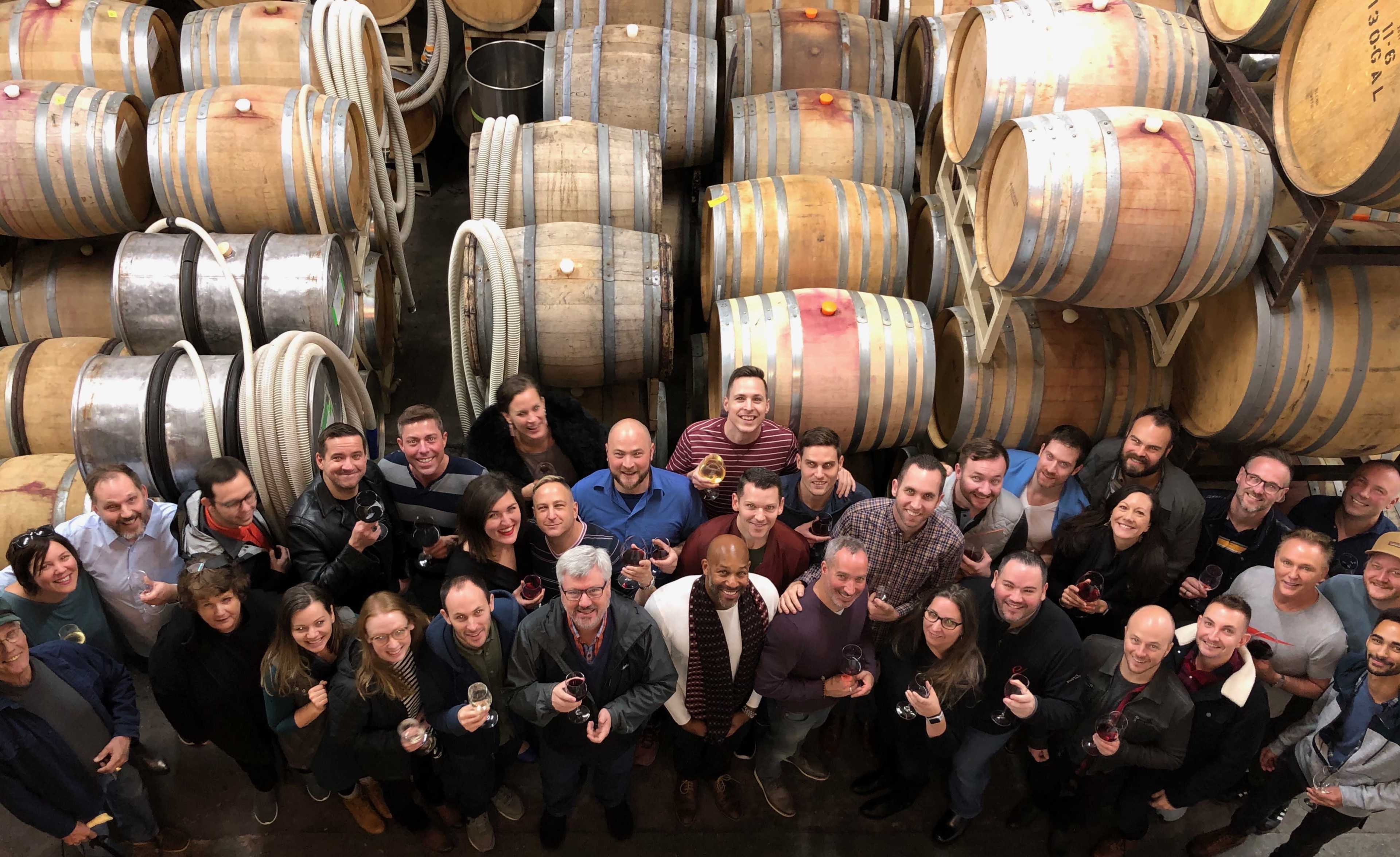 A large group of people stands together, holding glasses of wine, in a wine cellar surrounded by wooden barrels.