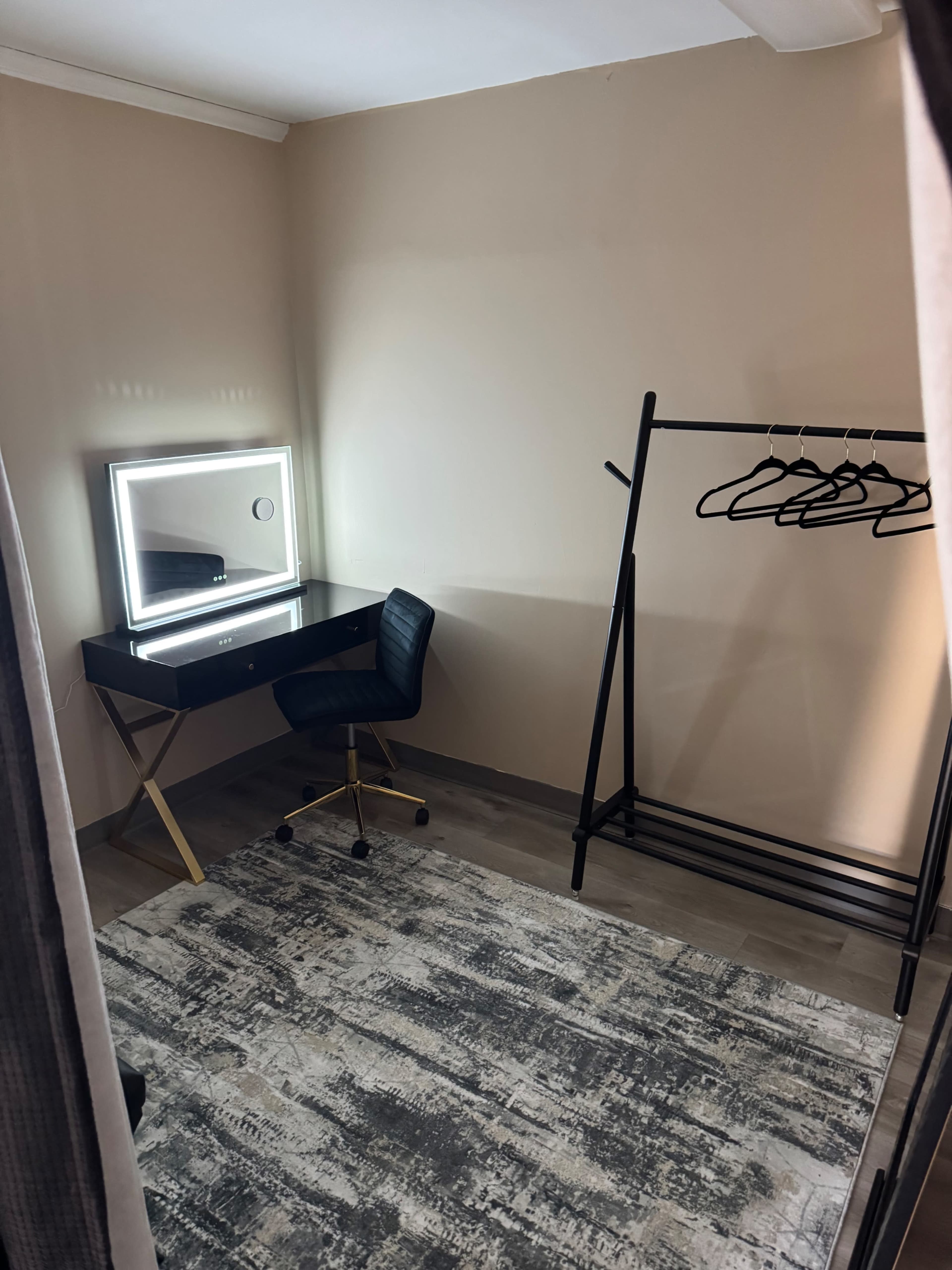 The image shows a minimalist room featuring a vanity mirror with lights, a desk chair, and a clothing rack with hangers against a neutral-colored wall.