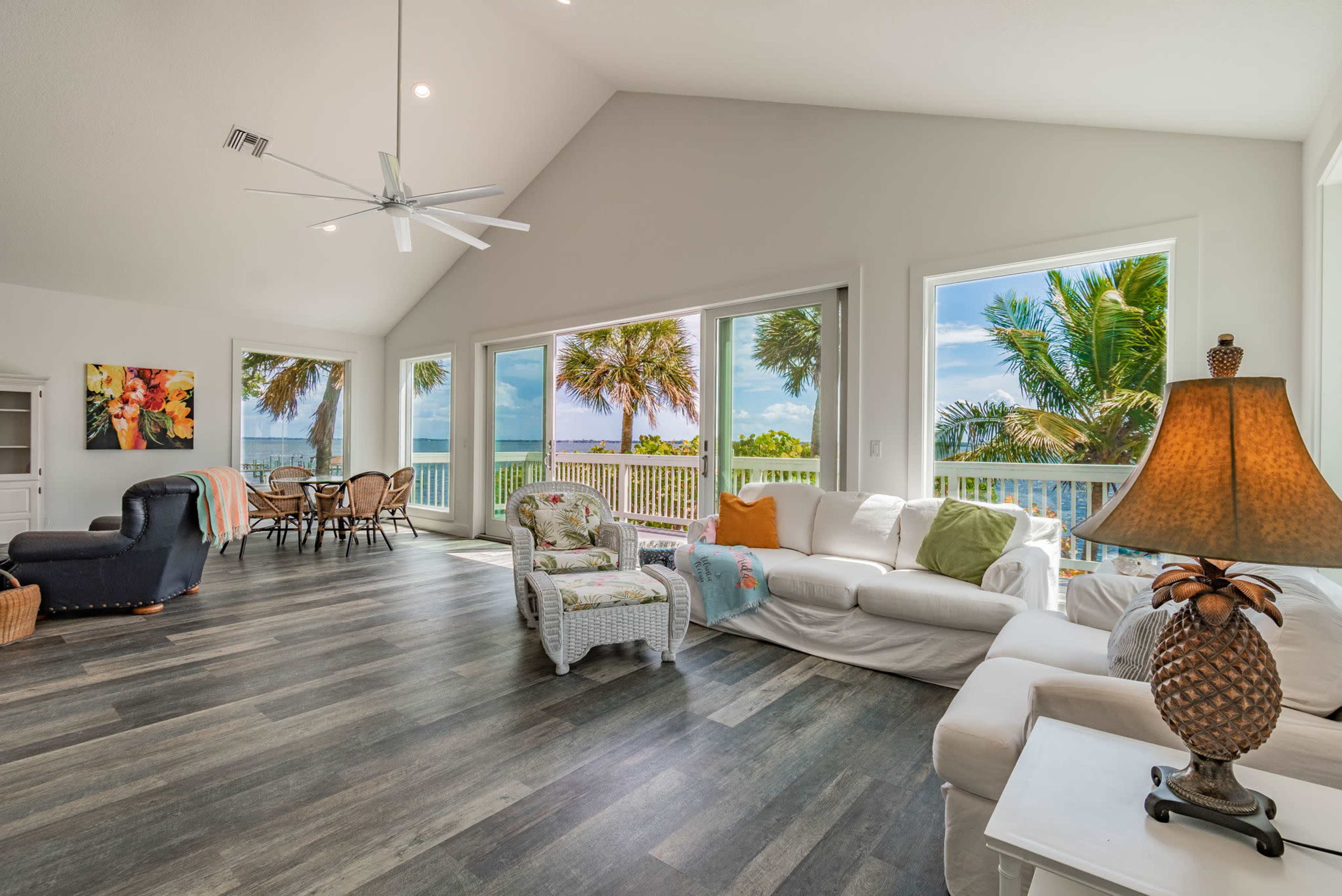 A spacious living room features large windows with views of palm trees and water, furnished with white sofas and a wicker chair.