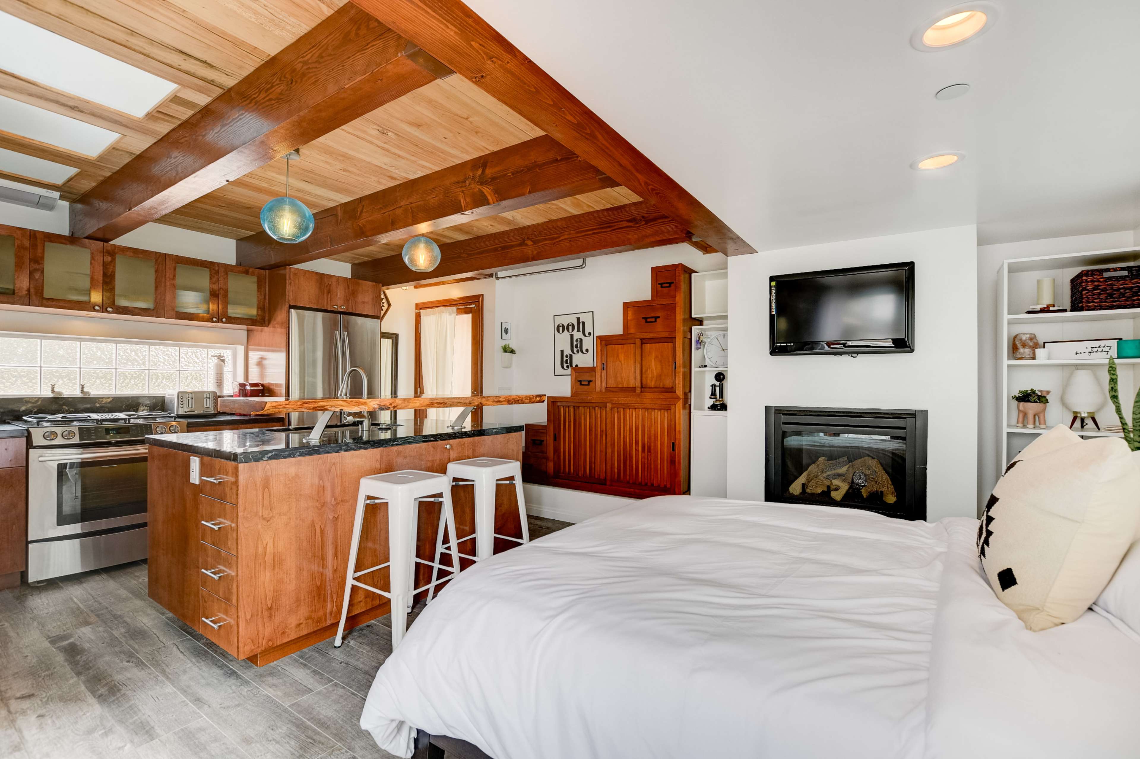A compact studio apartment featuring a kitchen with wooden beams, a small dining area with stools, a bed, and a fireplace.