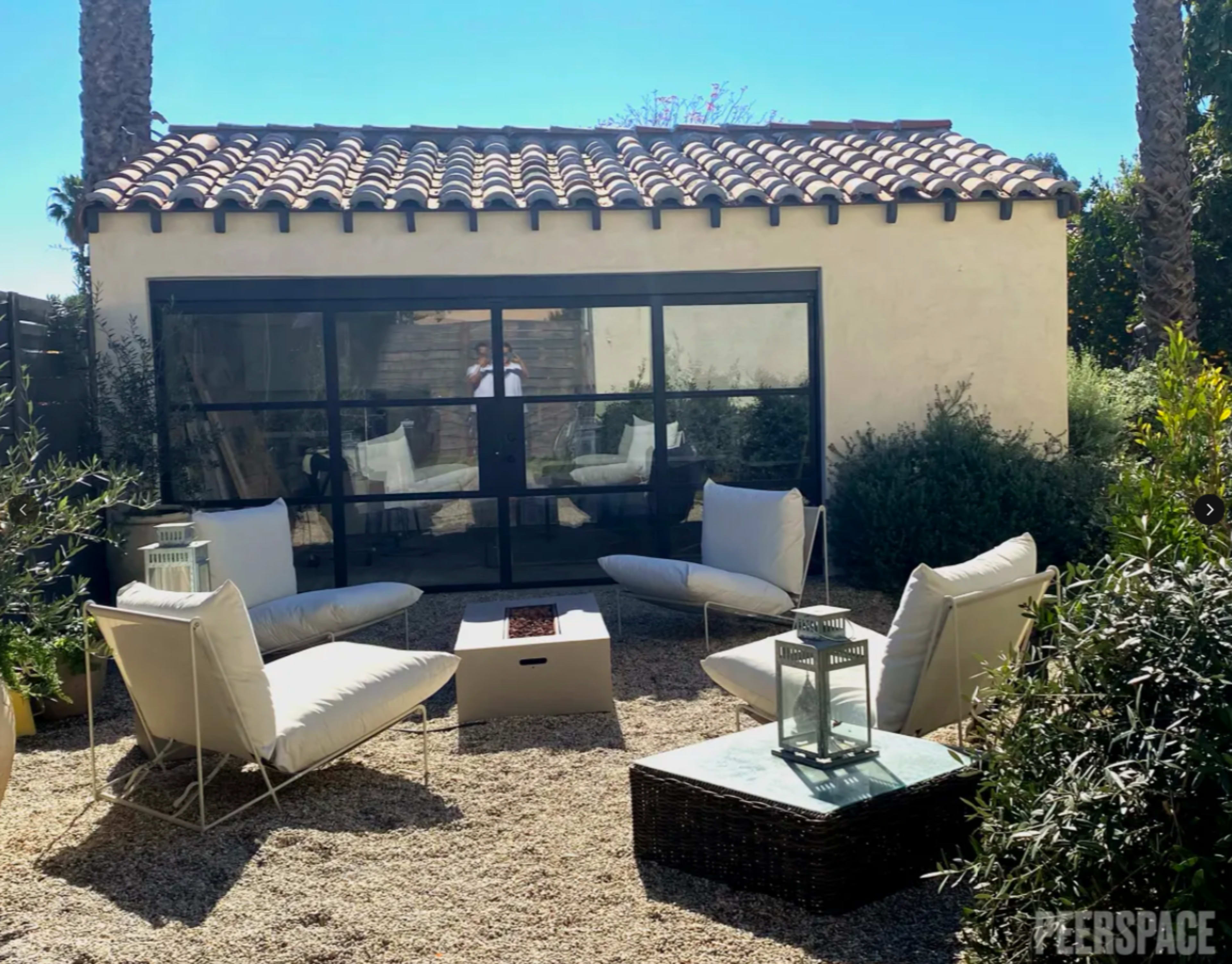 A cozy outdoor seating area features four white lounge chairs arranged around a fire pit, with a glass-walled structure in the background.