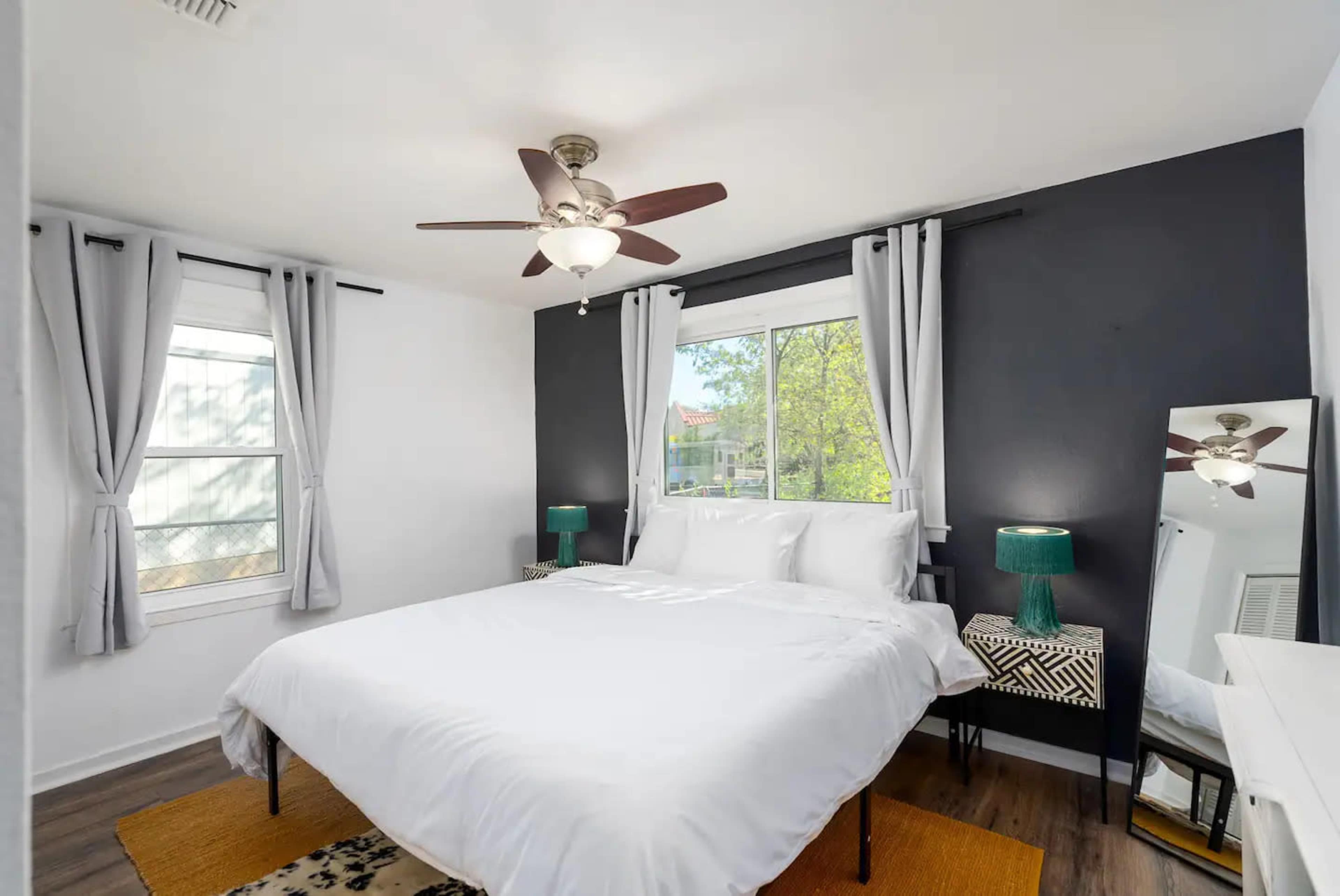 A well-lit bedroom features a large bed with white linens, two side tables with lamps, and a mirror beside a window with sheer curtains.