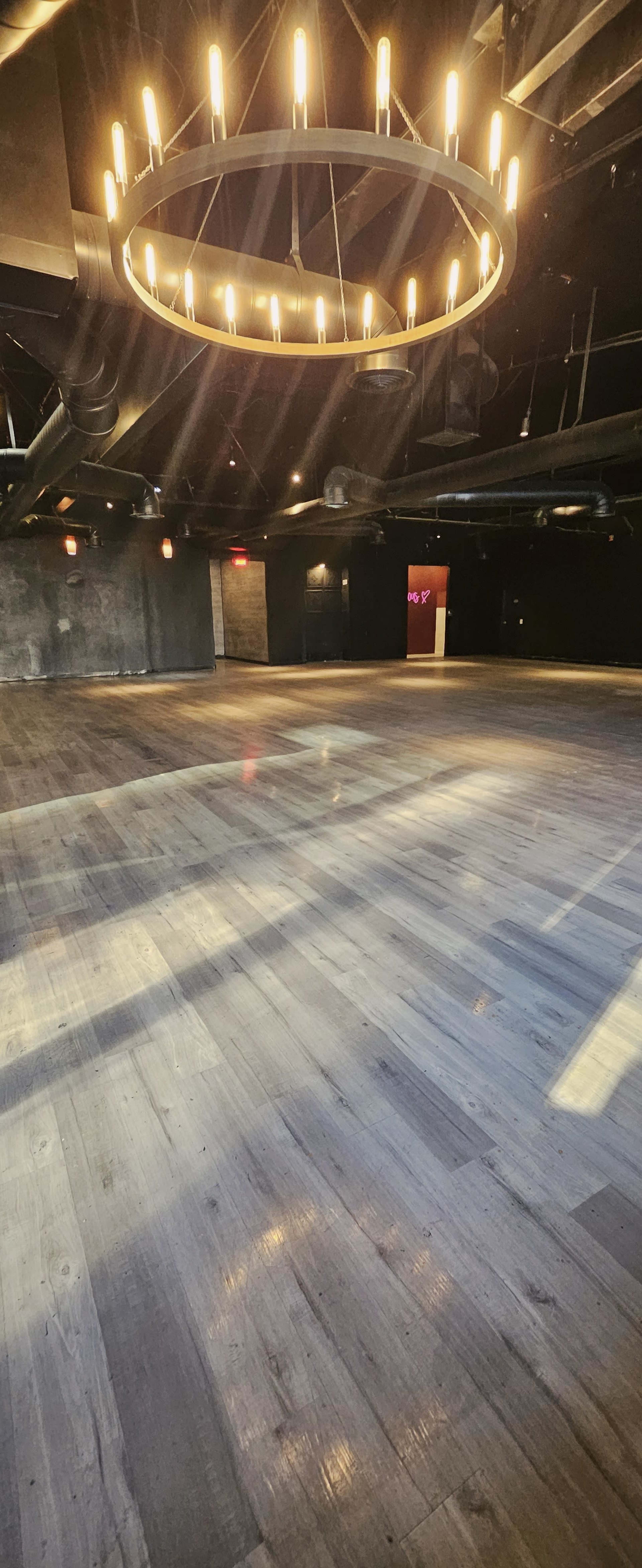 The image shows a spacious dimly lit room featuring a large circular chandelier and polished wooden flooring.