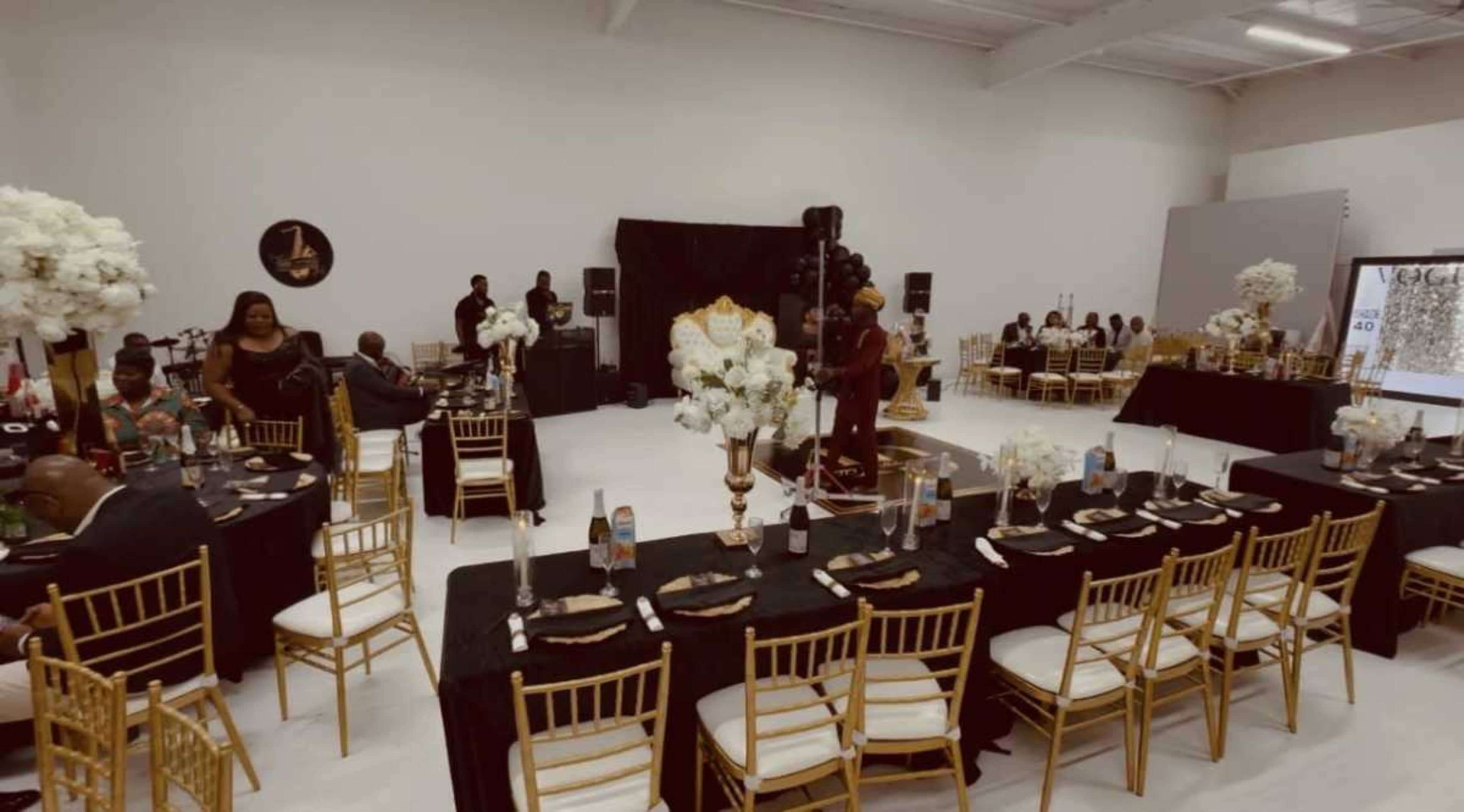 The image shows a spacious event venue decorated with black and gold tables, elegant floral centerpieces, and guests seated at round and long tables.