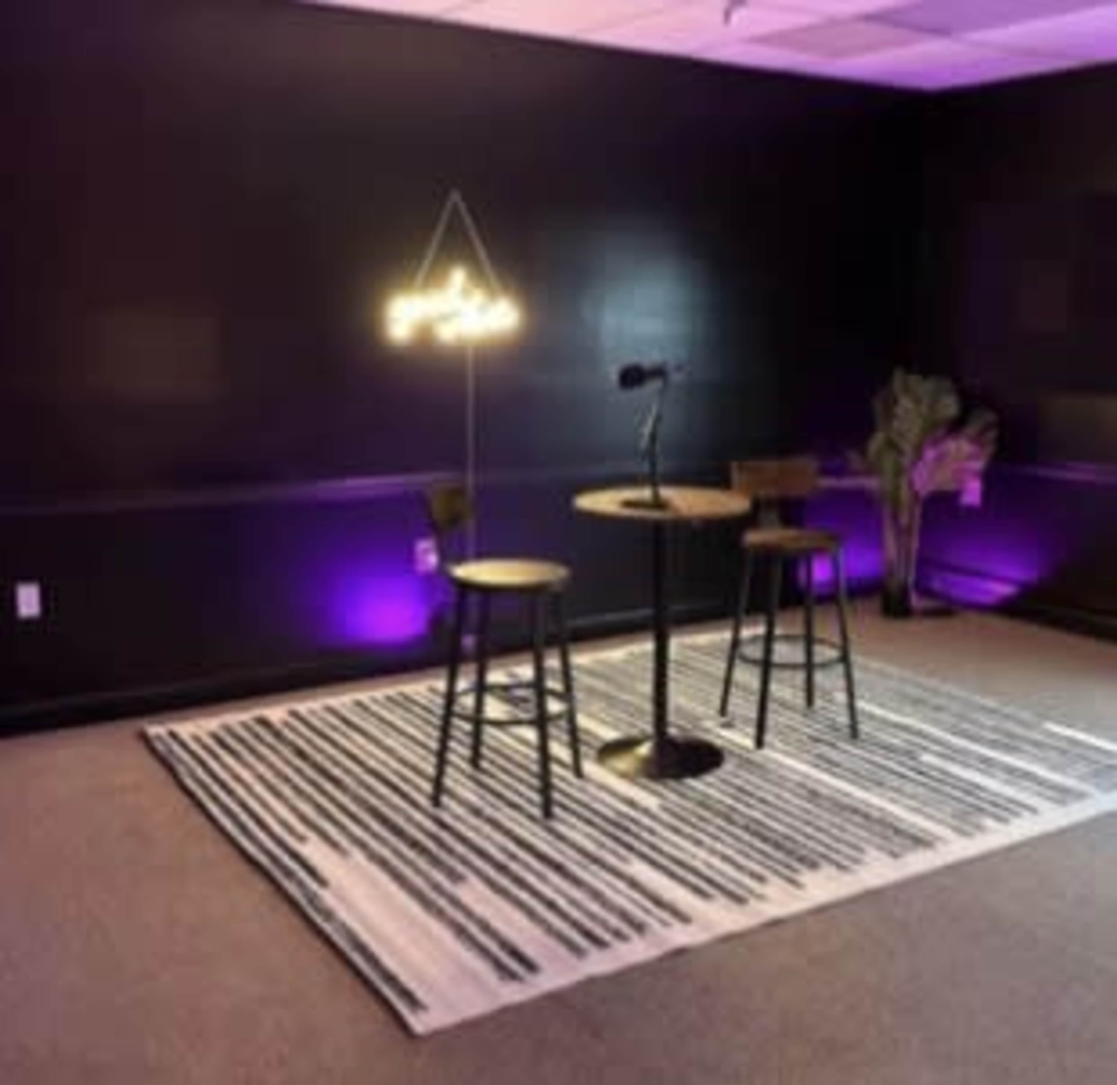 The image shows a small, modern room with two high stools, a round table, and a microphone, illuminated by purple lighting and a neon sign.