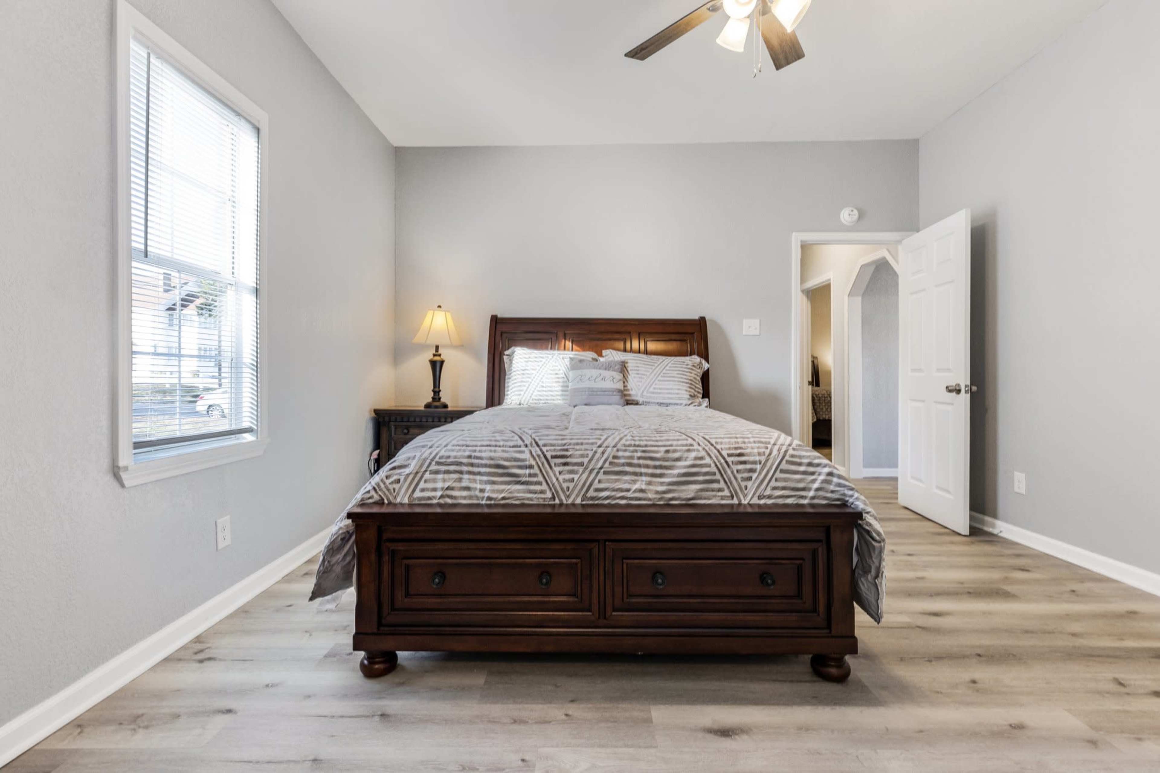 A neatly made bed with a striped comforter is centered in a well-lit bedroom featuring gray walls and a small bedside lamp.
