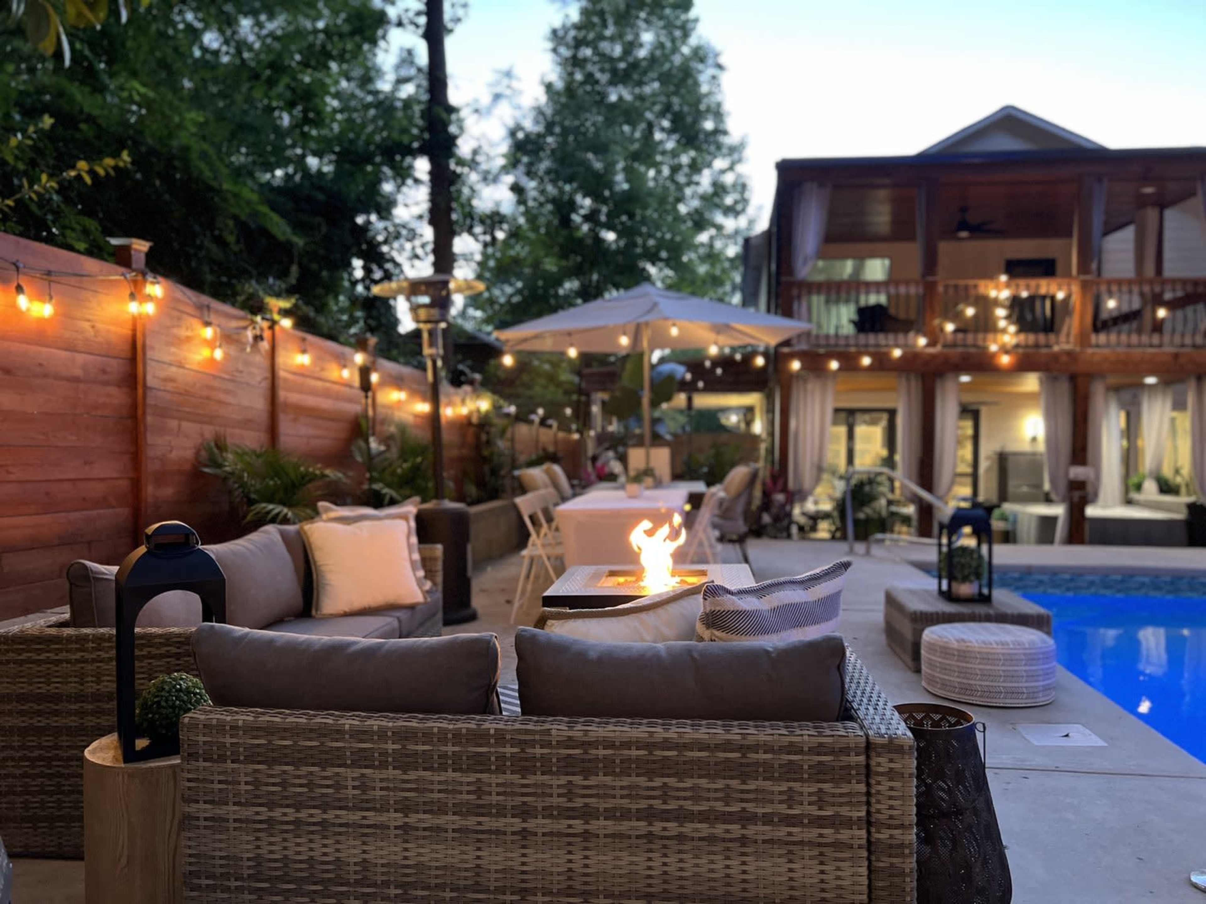 A cozy outdoor seating area with a fire pit, surrounded by a pool and string lights, in a backyard setting.