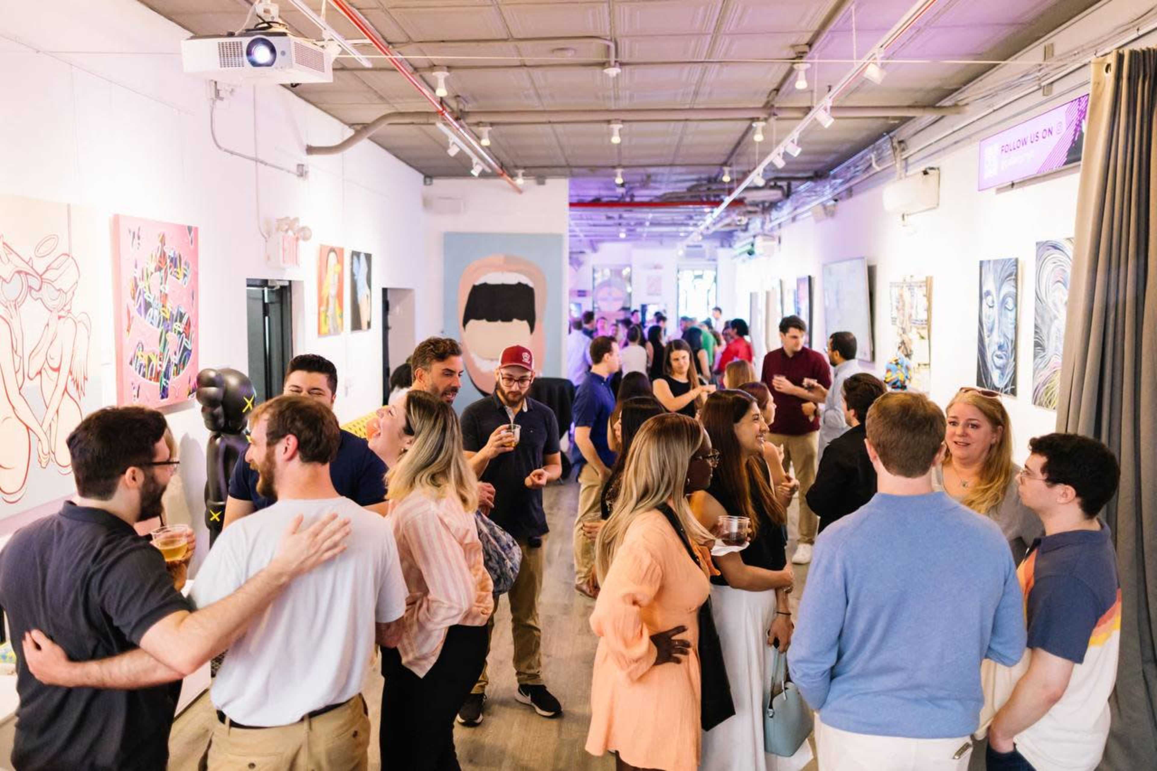 A group of people socialize at an art gallery opening featuring vibrant artworks on the walls.