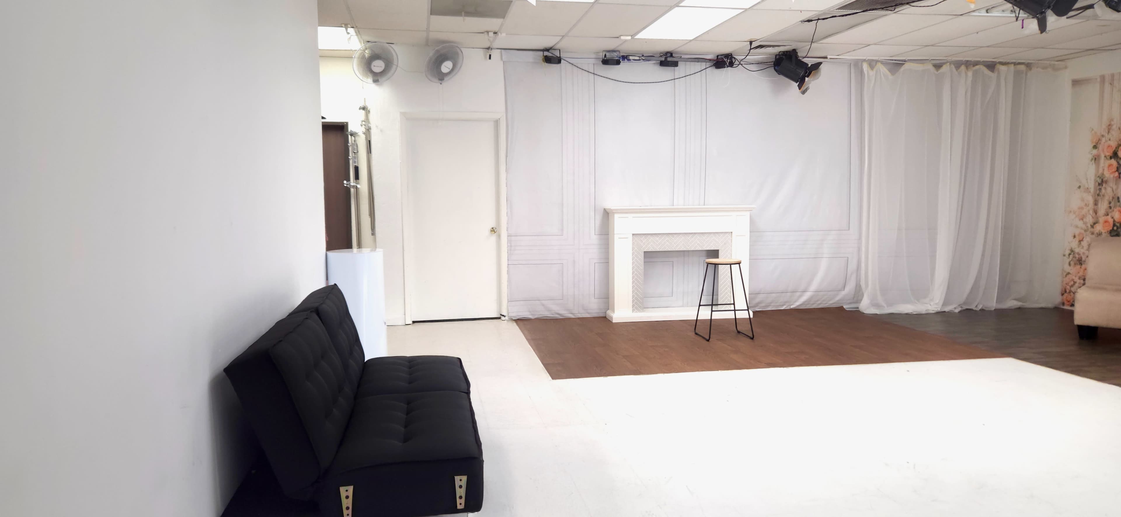 The image shows a spacious studio area with a black sofa, a wooden floor, a small stool in front of a white decorative fireplace, and light-colored walls.