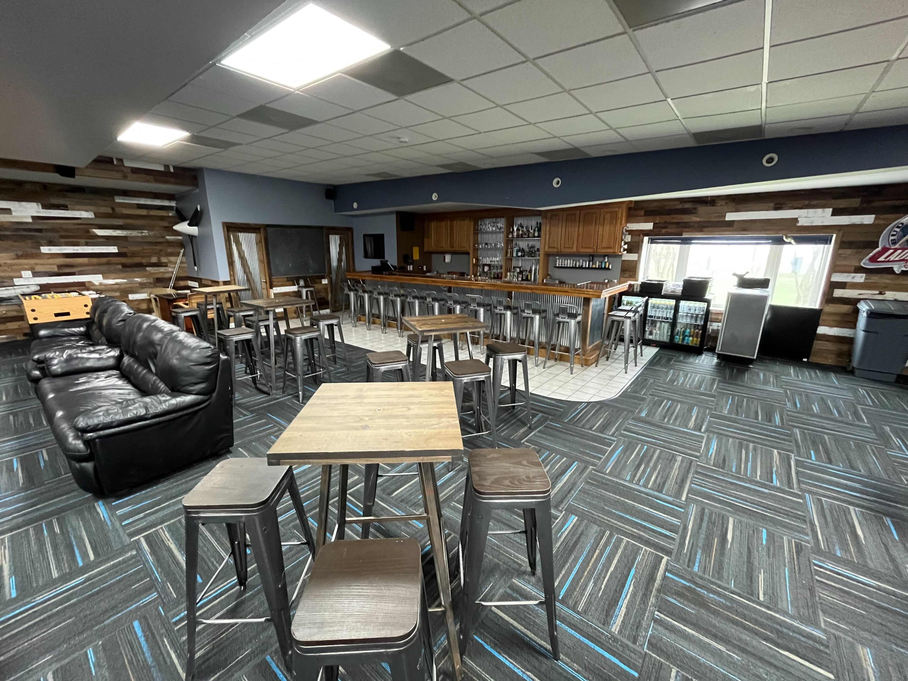 The image shows a spacious bar area with a central counter, high-top tables, stools, and a seating area equipped with a black leather couch.