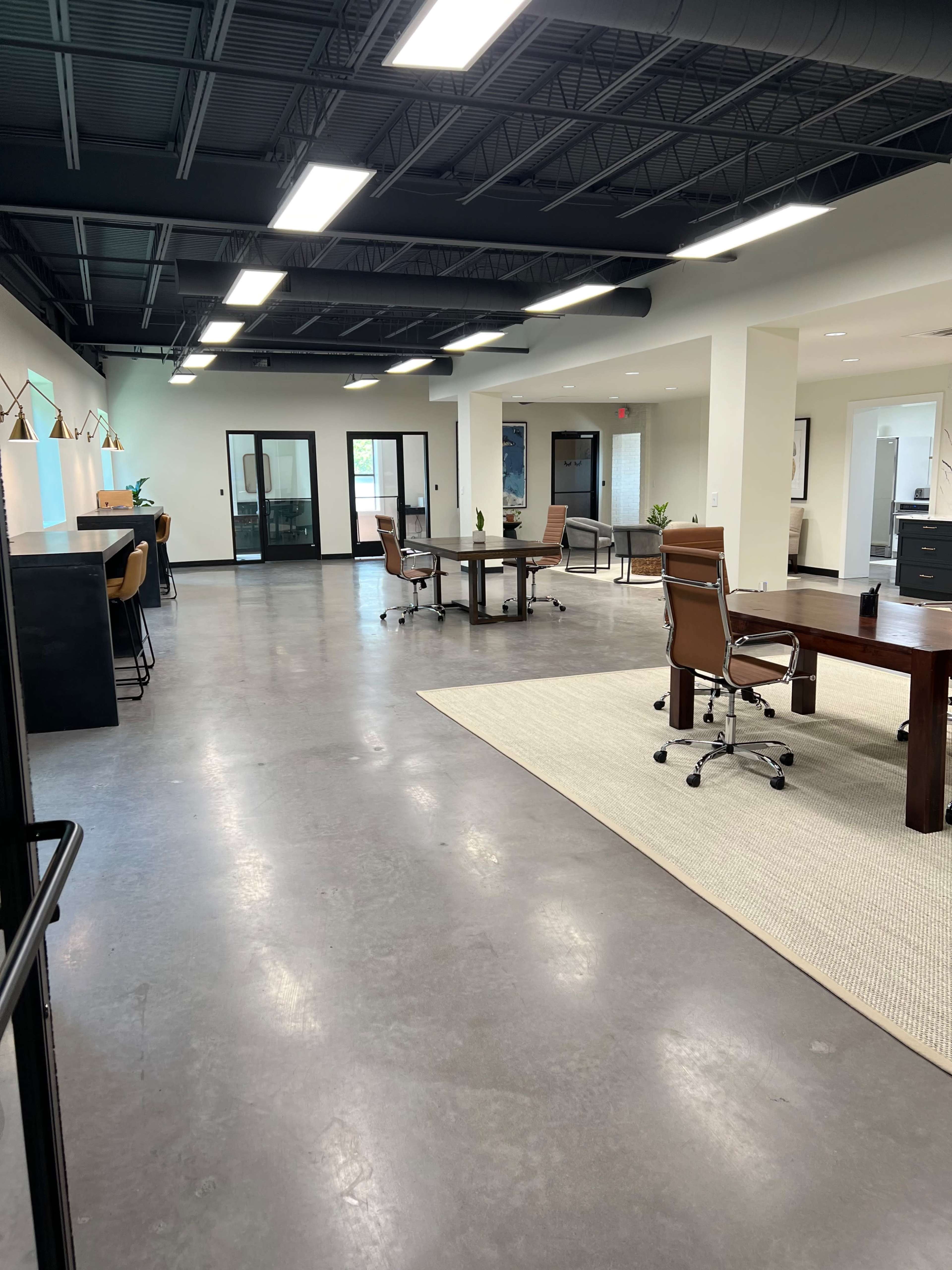 A spacious, modern office features multiple workstations, including desks and chairs, with concrete flooring and large windows.