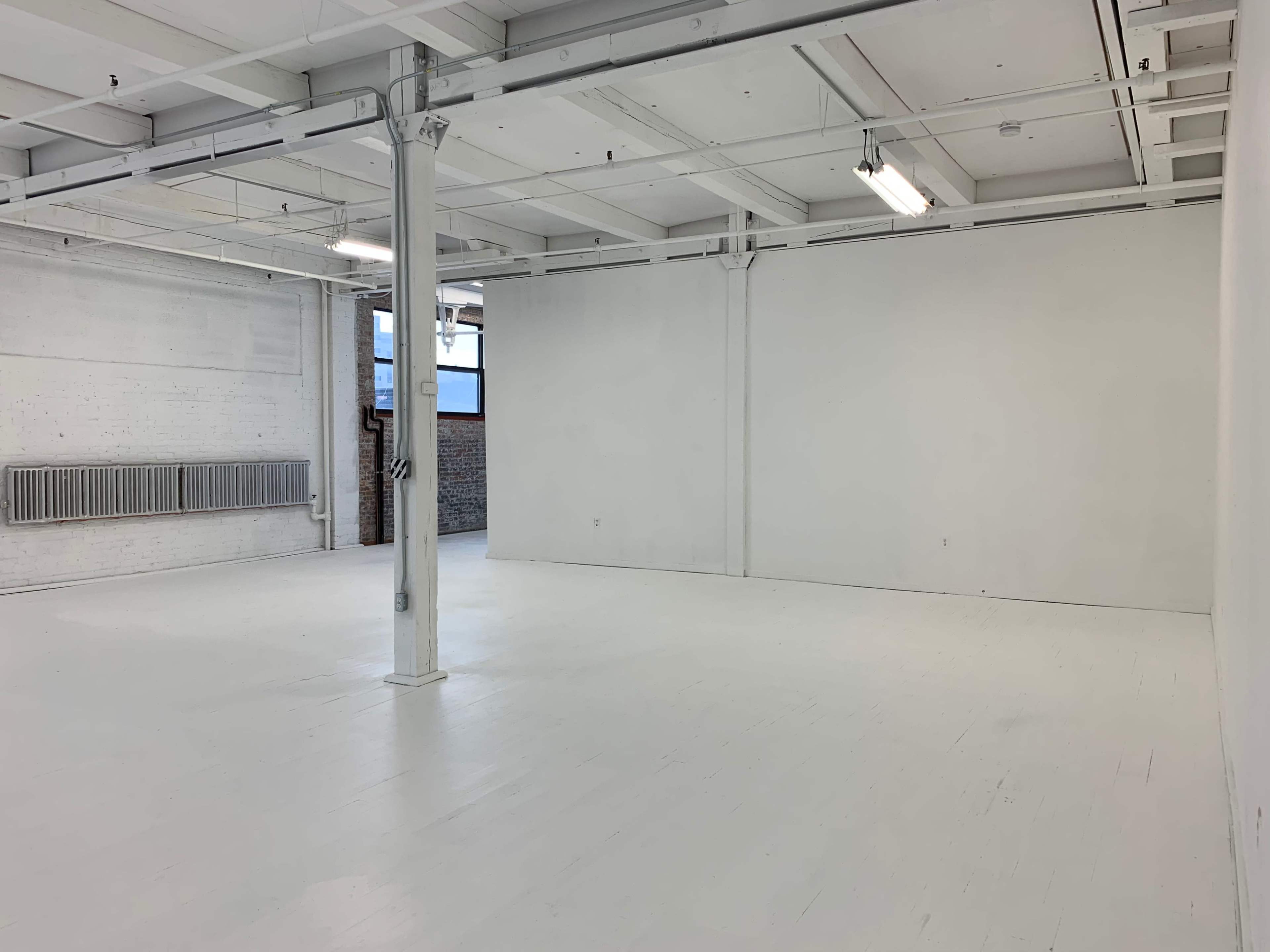 The image shows a large, empty industrial-style room with white walls and a polished floor, featuring a central support column and large windows allowing natural light to enter.