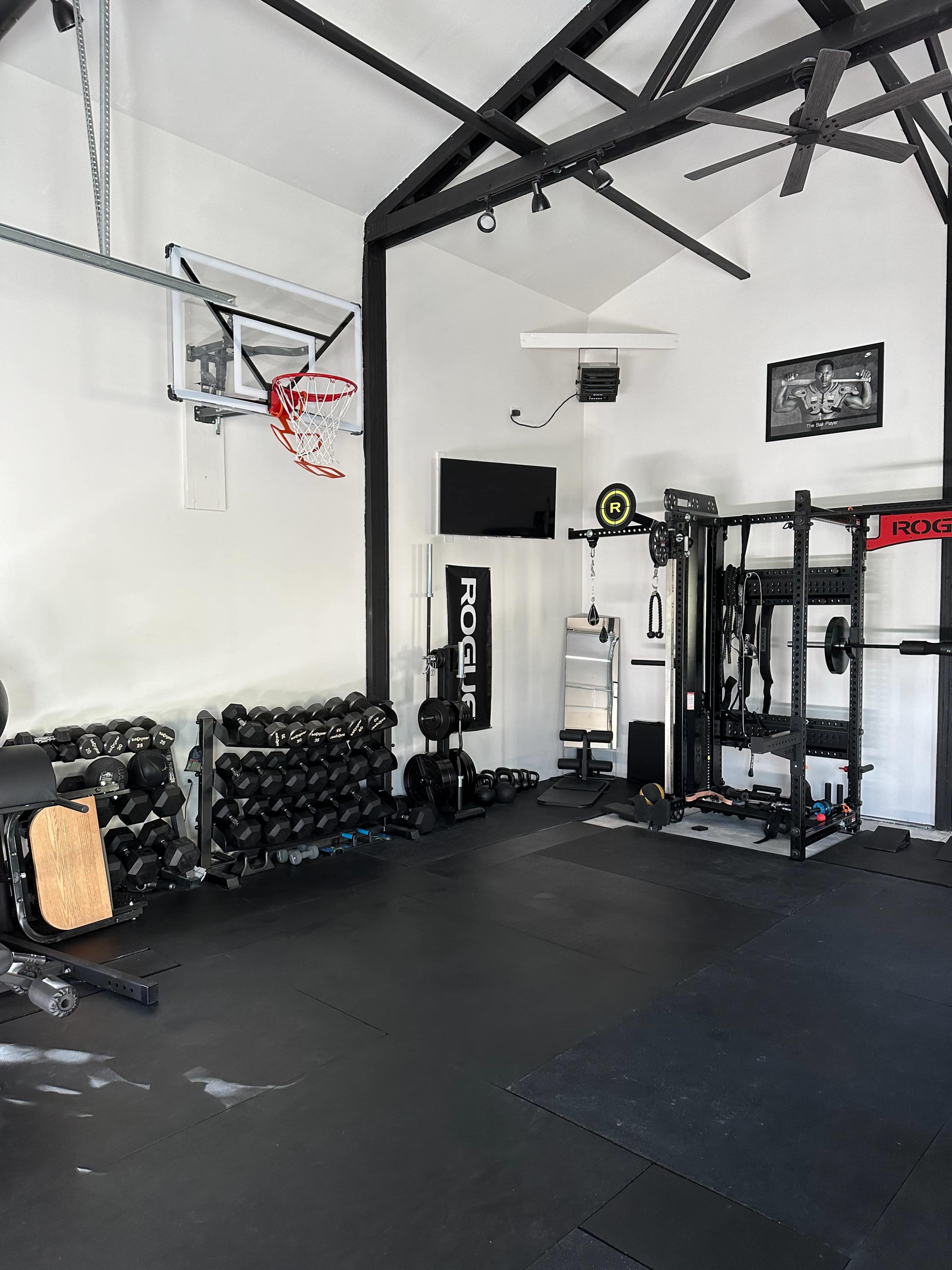 The image shows an indoor gym featuring a variety of fitness equipment, including dumbbells, a weight rack, a cable machine, and a wall-mounted basketball hoop.