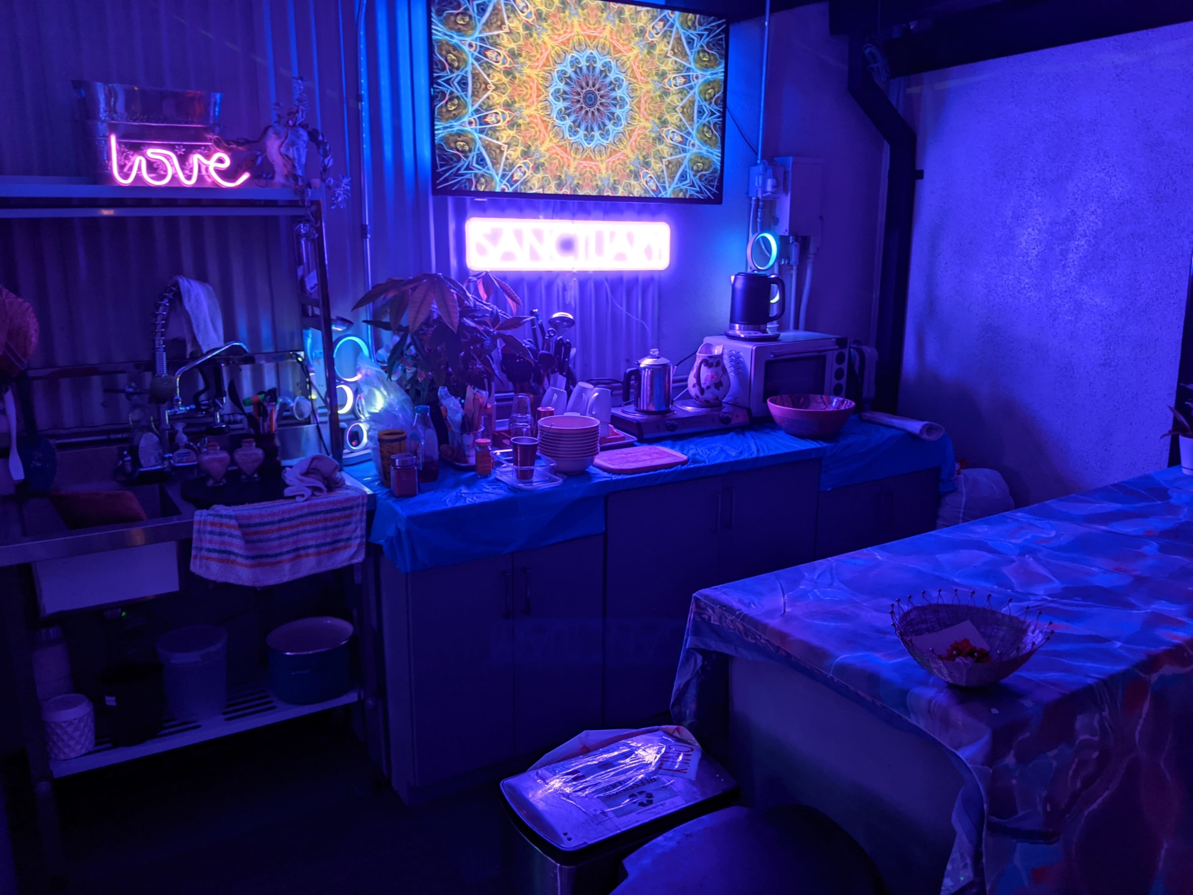 The image shows a colorful kitchen space with a neon sign, a large decorative display on a screen, and various kitchen appliances and utensils arranged around a table.