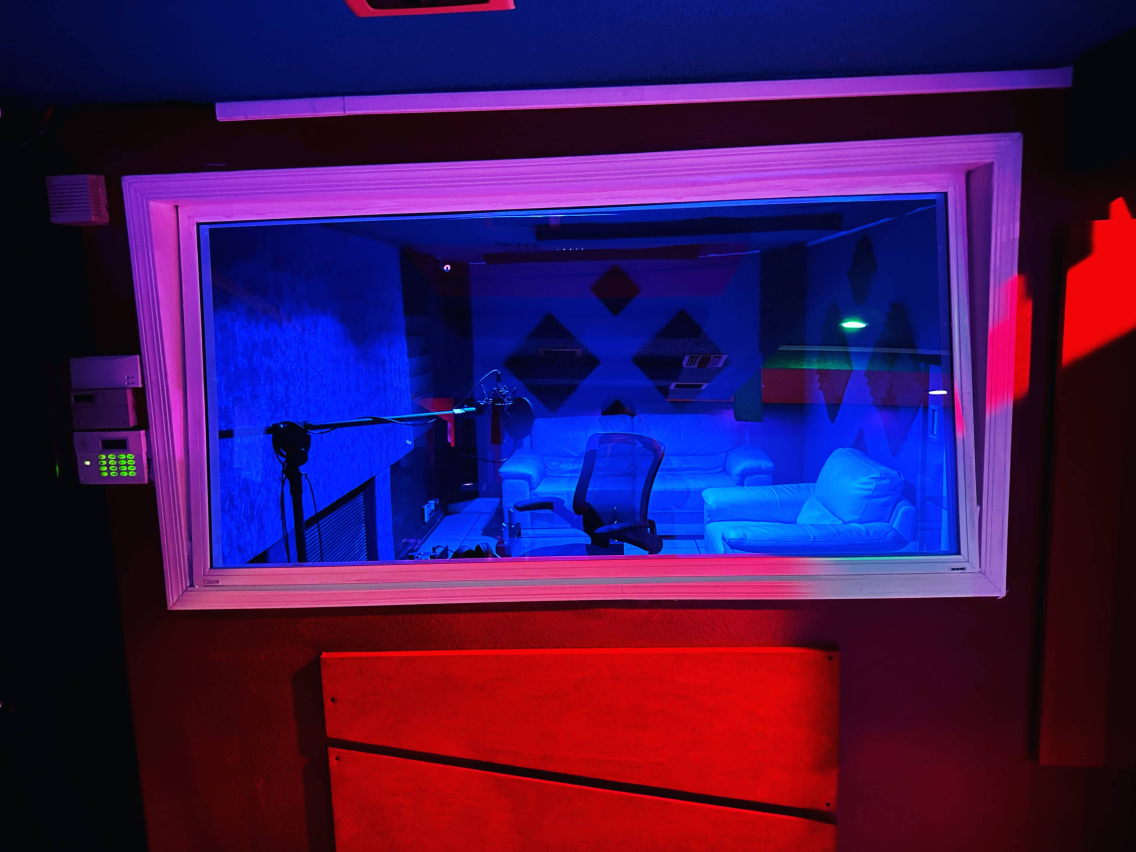 The image shows a recording studio with a glass window, featuring a microphone, a chair, and white couches illuminated by blue and red lighting.
