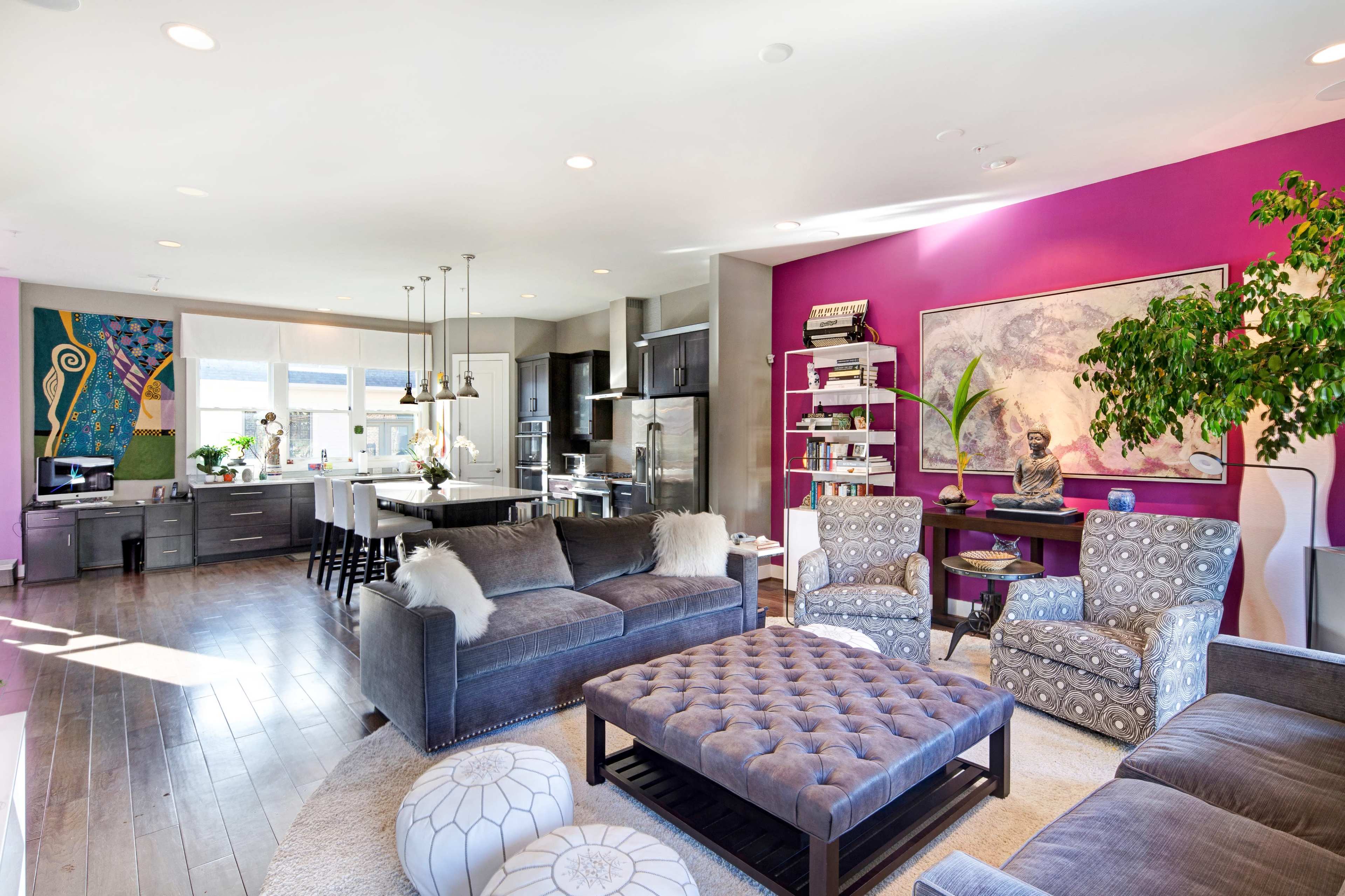 The image shows a contemporary living room featuring a plush gray sofa, patterned armchairs, a tufted coffee table, and bold purple accent walls, with a dining area in the background.