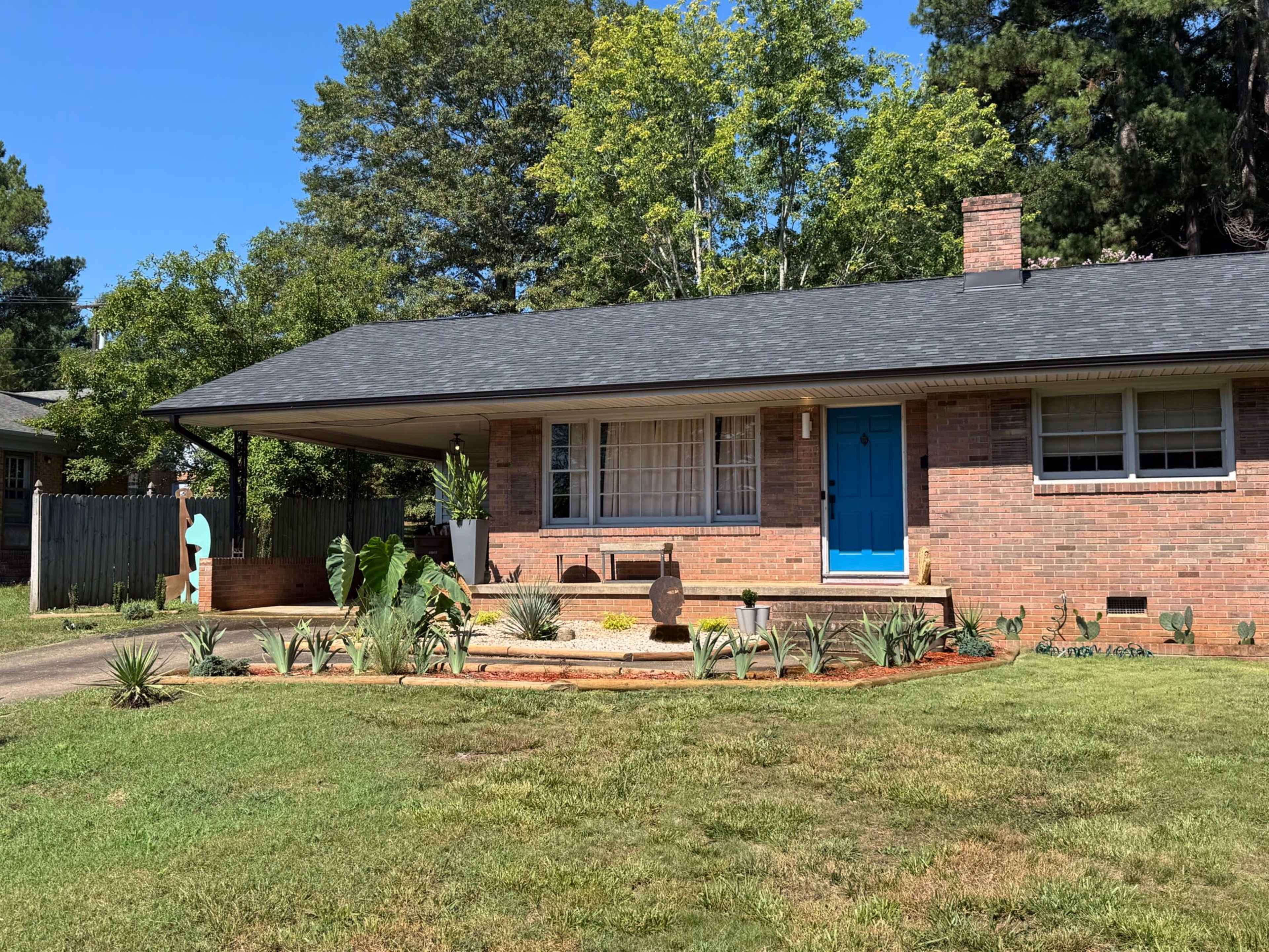 The image shows a single-story brick house with a blue front door, surrounded by a landscaped yard featuring plants and decorative stones.