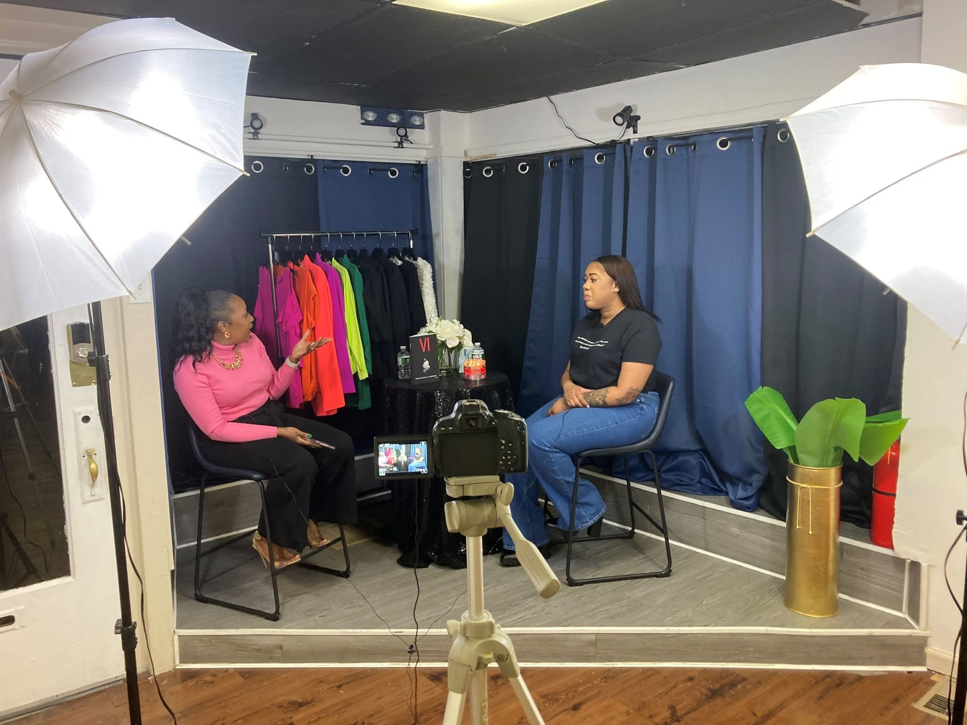 Two women are seated in a studio setting for an interview, with a rack of colorful clothing visible in the background.
