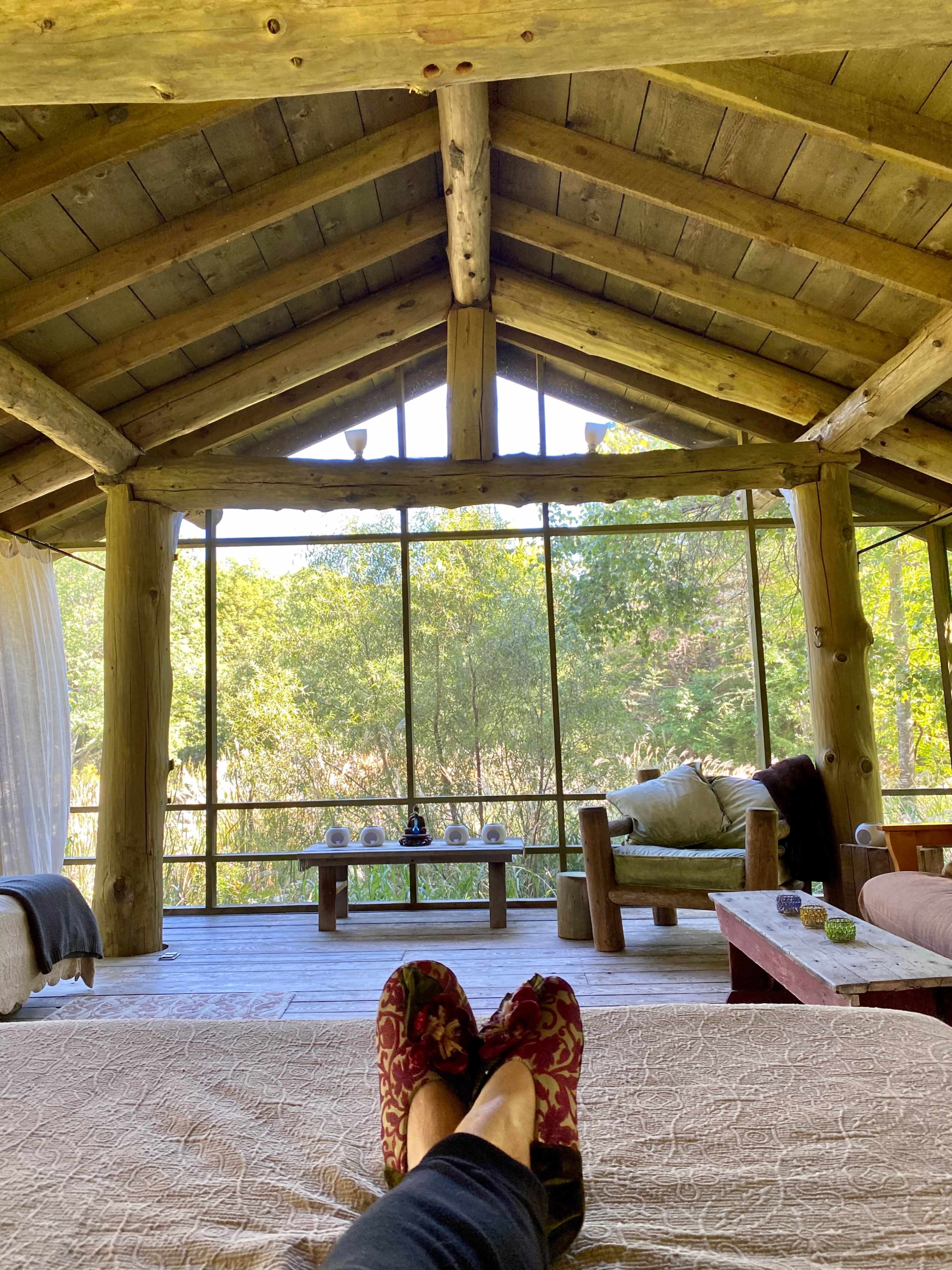 A person relaxes on a bed inside a rustic cabin, with large windows offering a view of greenery outside.