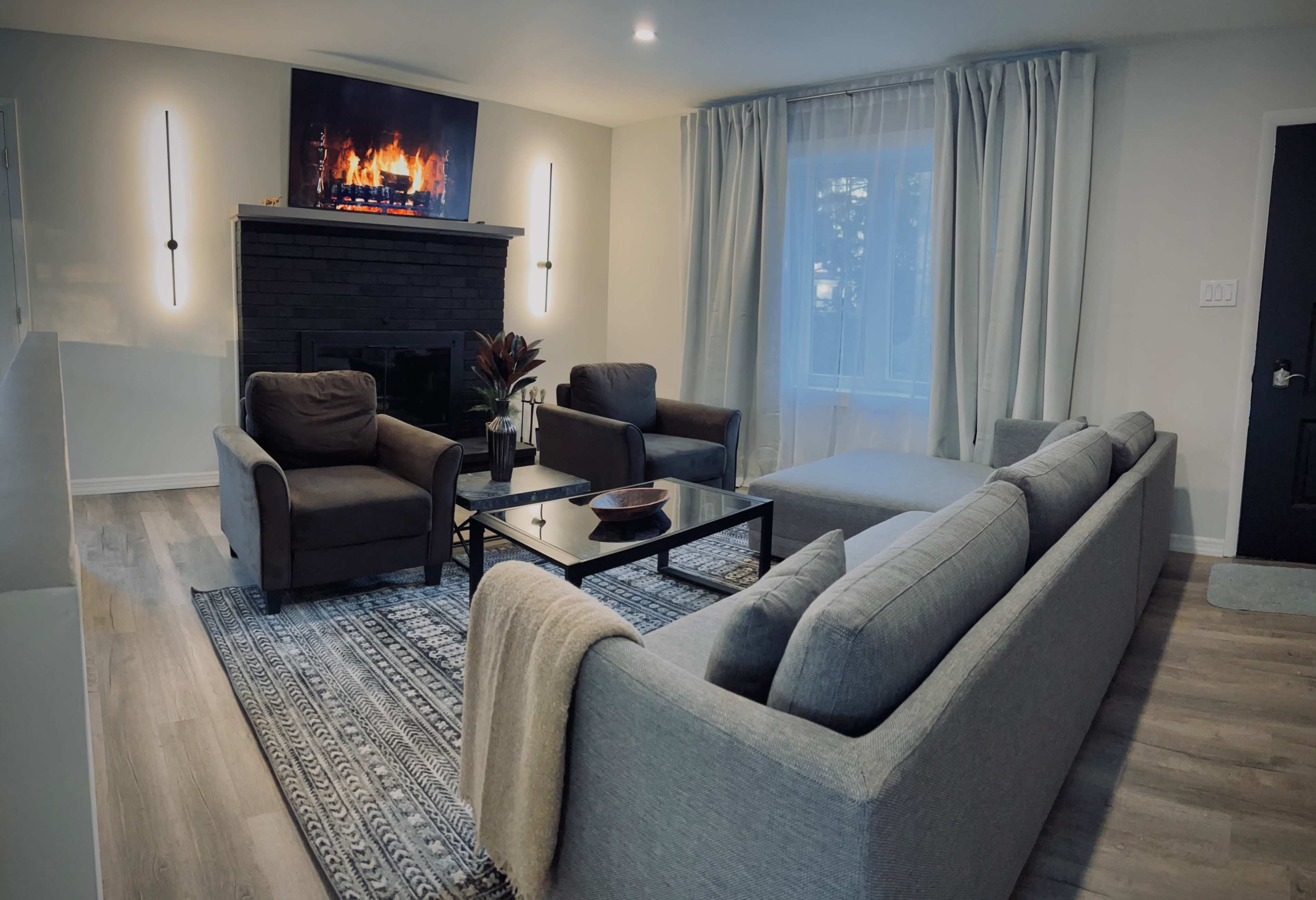 A cozy living room with a gray sofa, two armchairs, a coffee table, and a wall-mounted television displaying a fireplace.