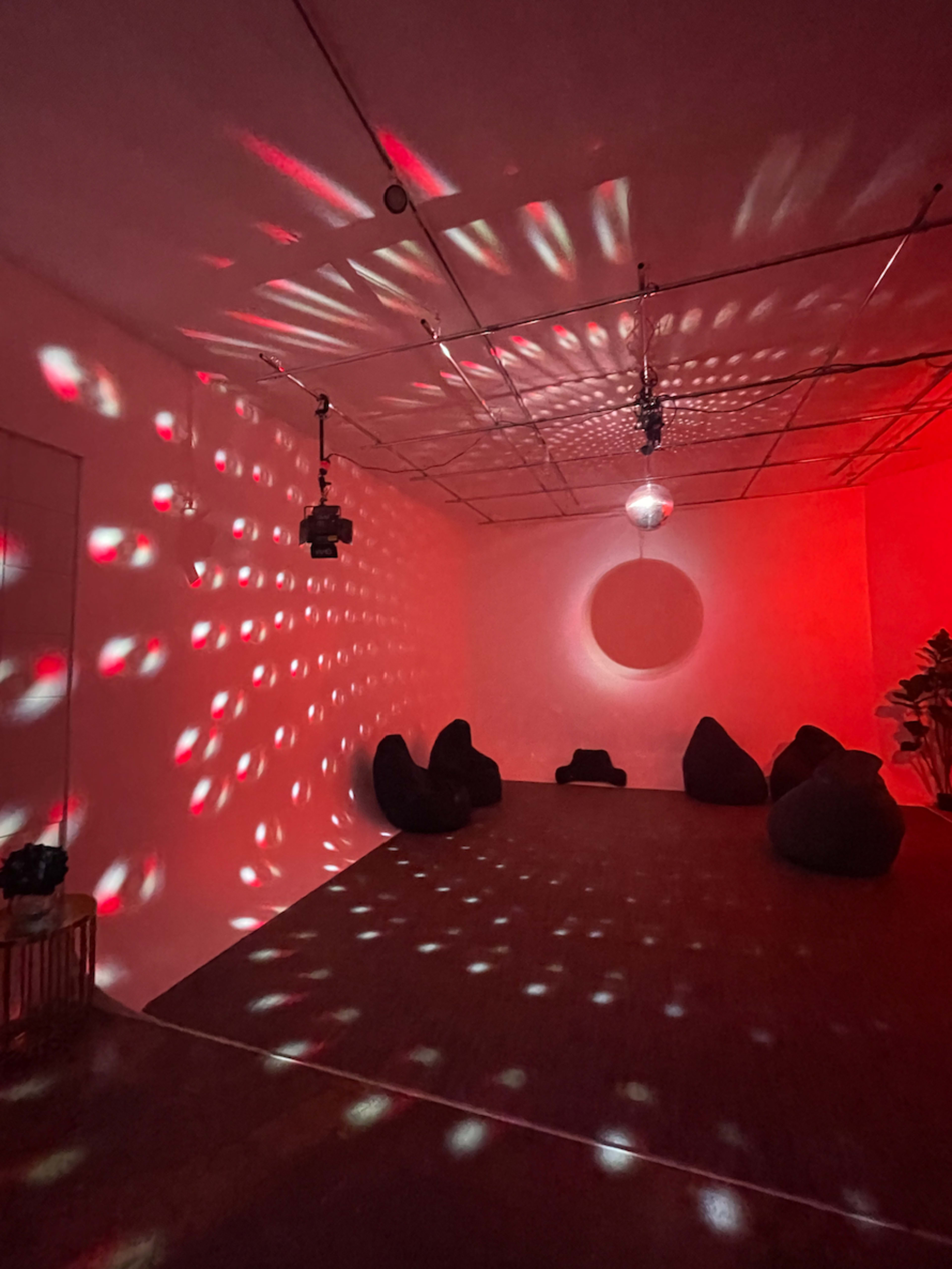 A room with a disco ball casting colorful light patterns on the walls, featuring several black lounge cushions and a circular wall decoration.