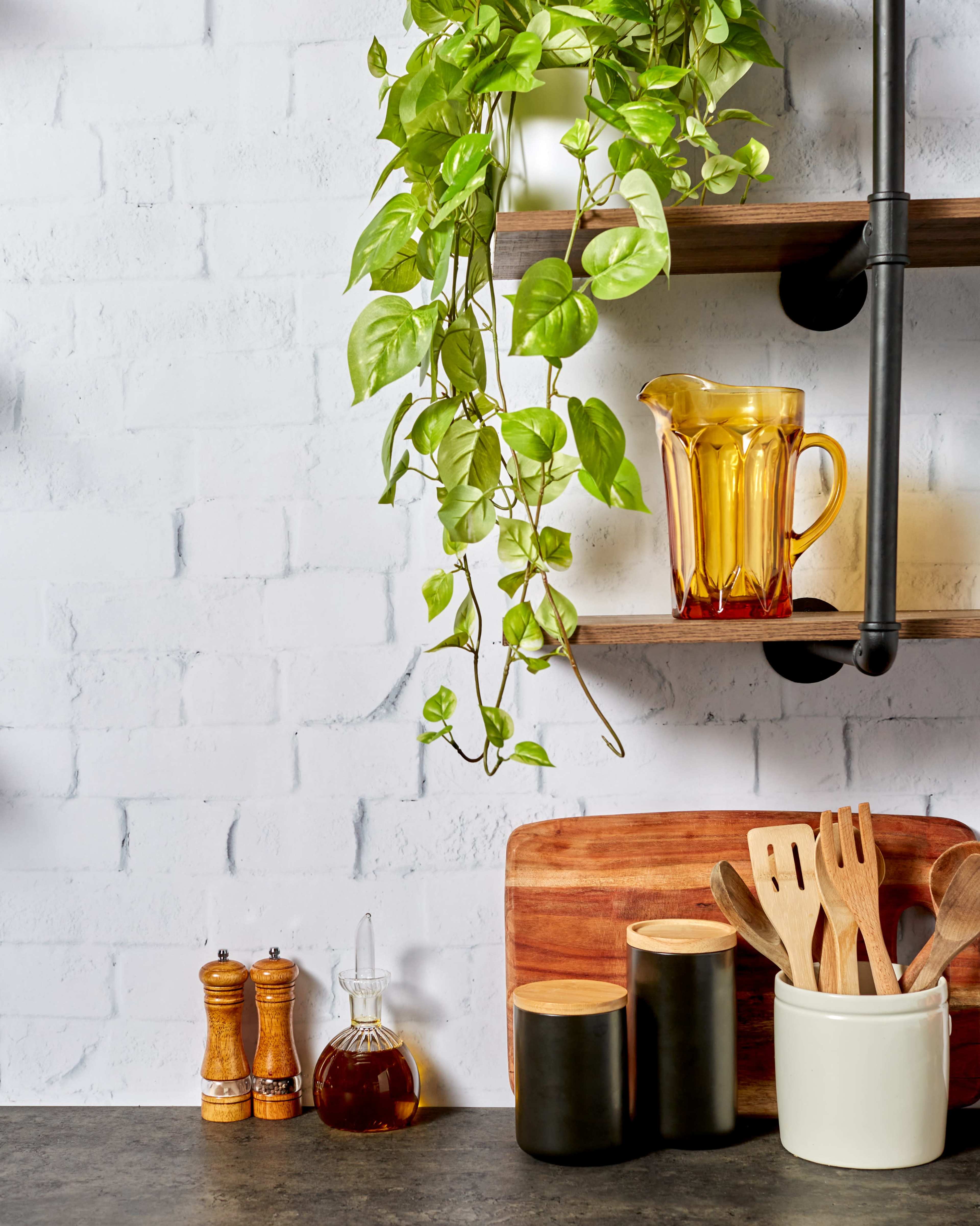 A kitchen countertop features wooden utensils in a container, a glass pitcher, and decorative greenery against a brick wall.
