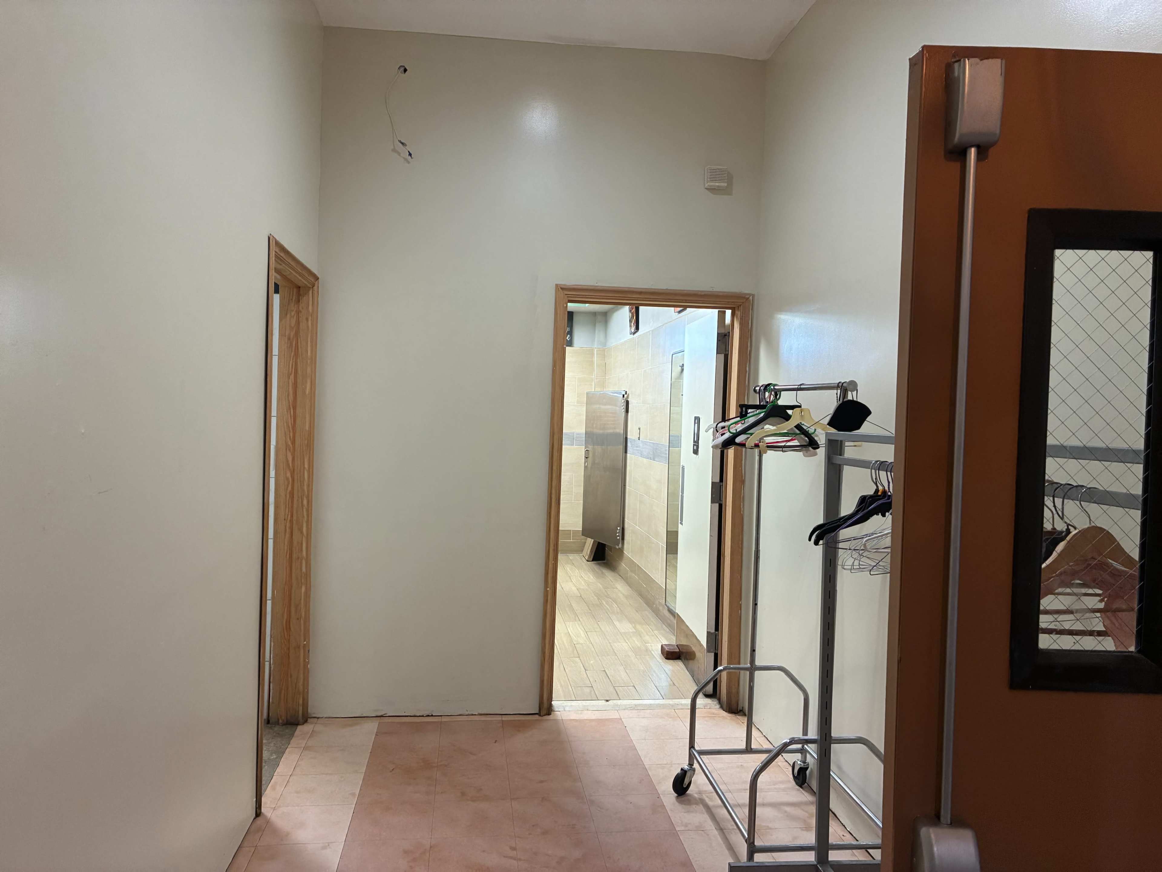 The image shows a hallway leading to a bathroom, with a clothing rack on the left side and a doorframe that opens to a tiled area.