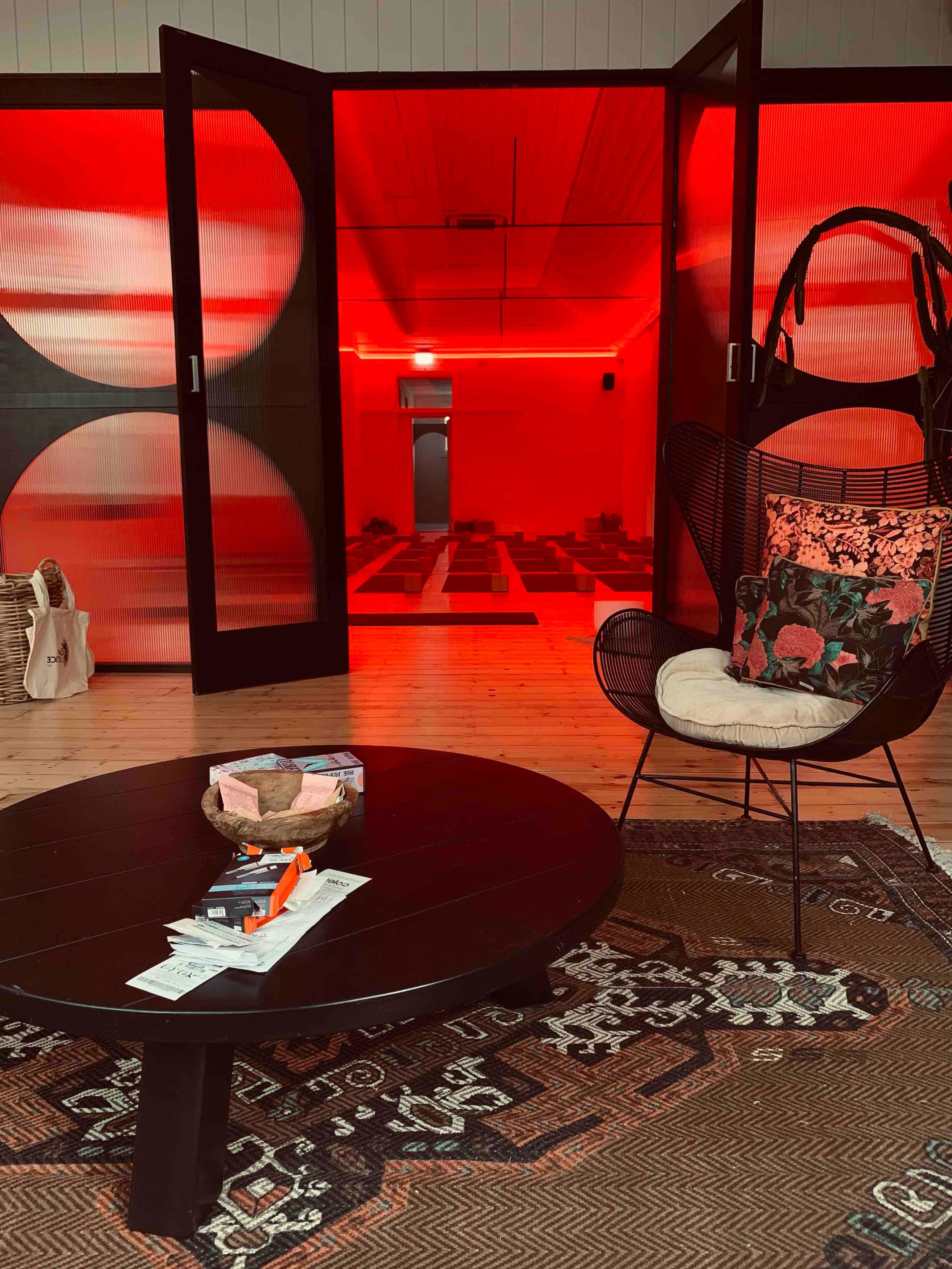 A modern interior features a round black table with magazines and a decorative bowl, surrounded by warm red lighting highlighting an open doorway and a hanging chair.