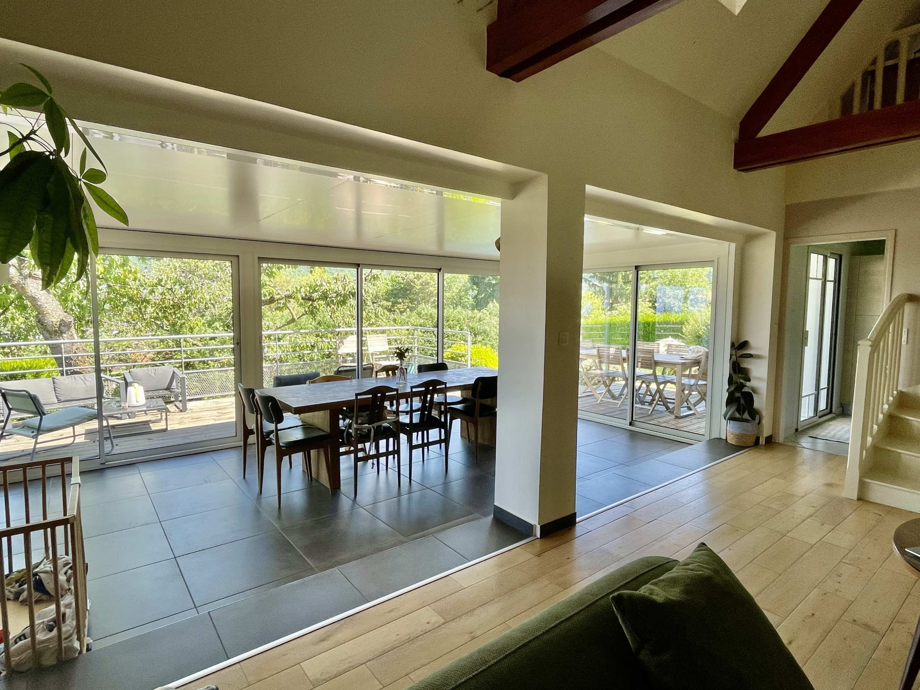 The image shows a modern, spacious interior with large glass doors opening to an outdoor area, featuring a dining table and chairs inside, along with a green couch and a staircase visible in the background.