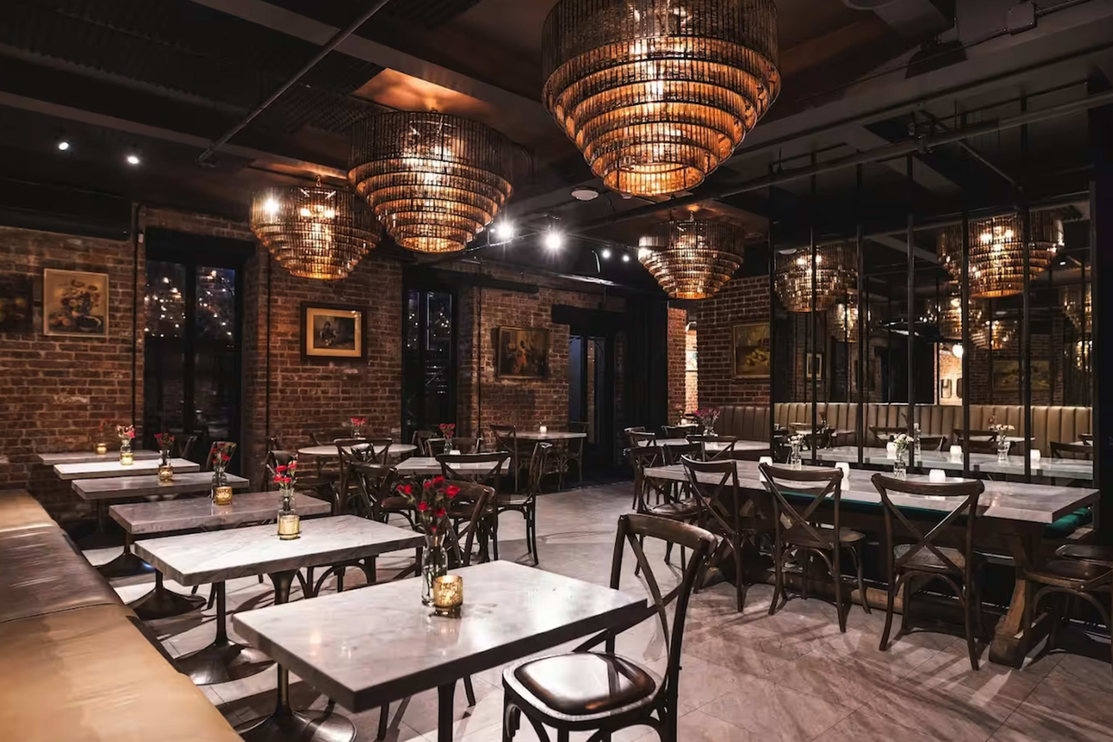 The image shows a stylish restaurant interior featuring wooden tables, chairs, and large woven light fixtures overhead, with red rose centerpieces on each table.
