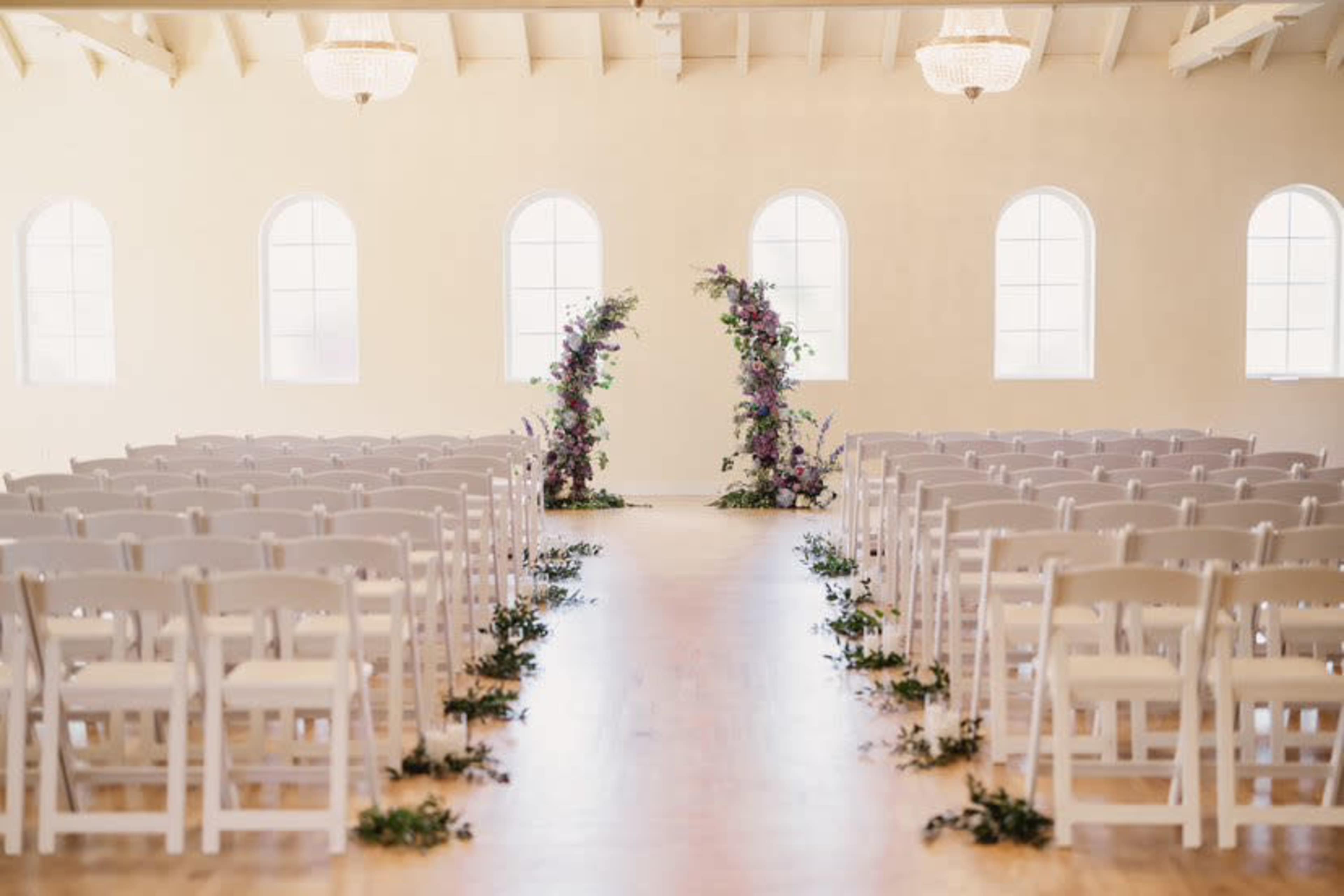 The image shows a wedding ceremony setup featuring rows of white chairs facing an aisle adorned with flowers and two floral arches at the end.