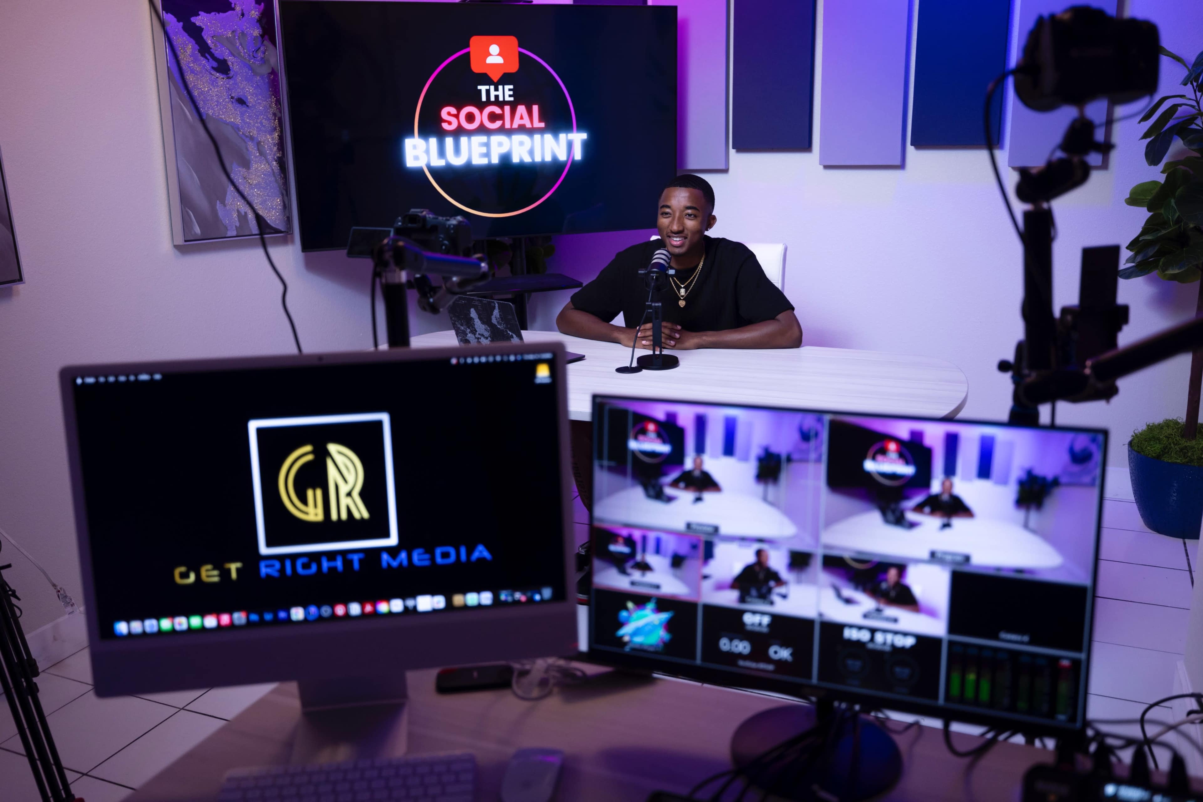 A man sits at a desk with a microphone in front of him, surrounded by multiple screens displaying a logo and video feeds in a well-lit studio setting.