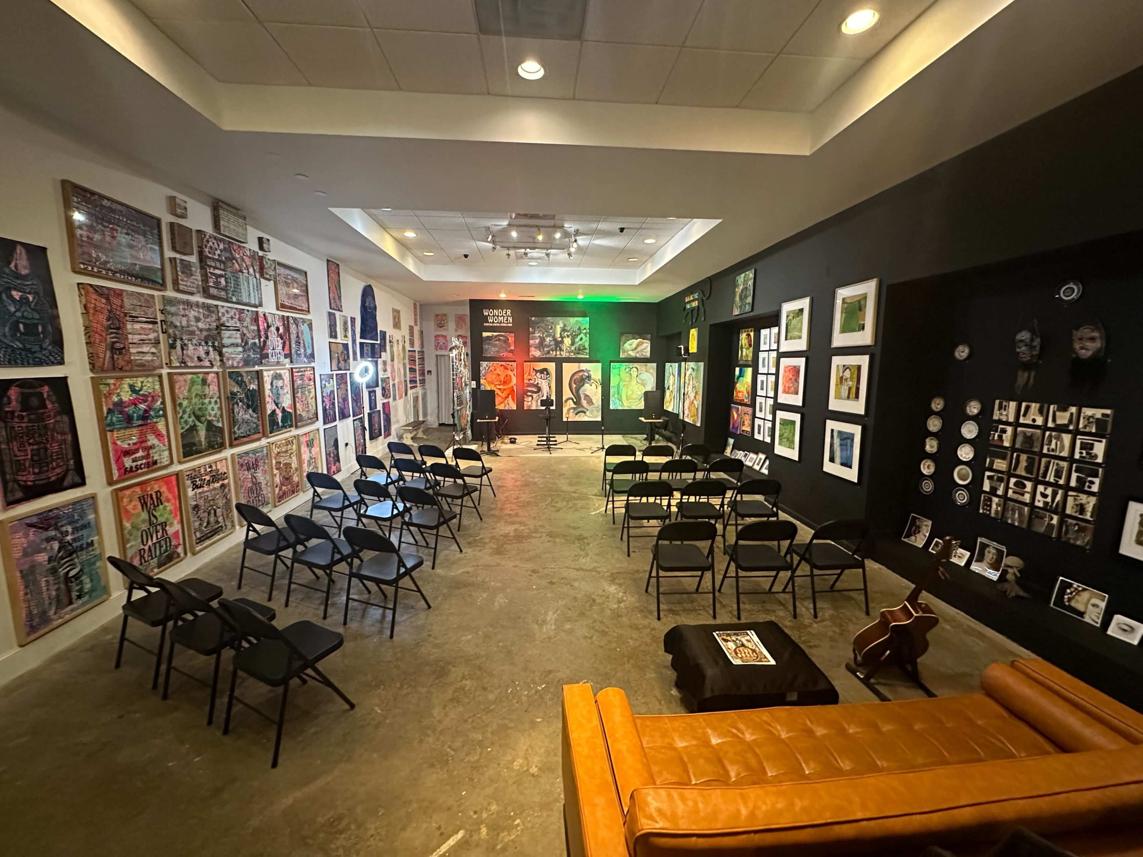 The image shows an art gallery featuring walls covered in various artworks, with rows of black folding chairs arranged in front and a lounge area with a brown sofa.