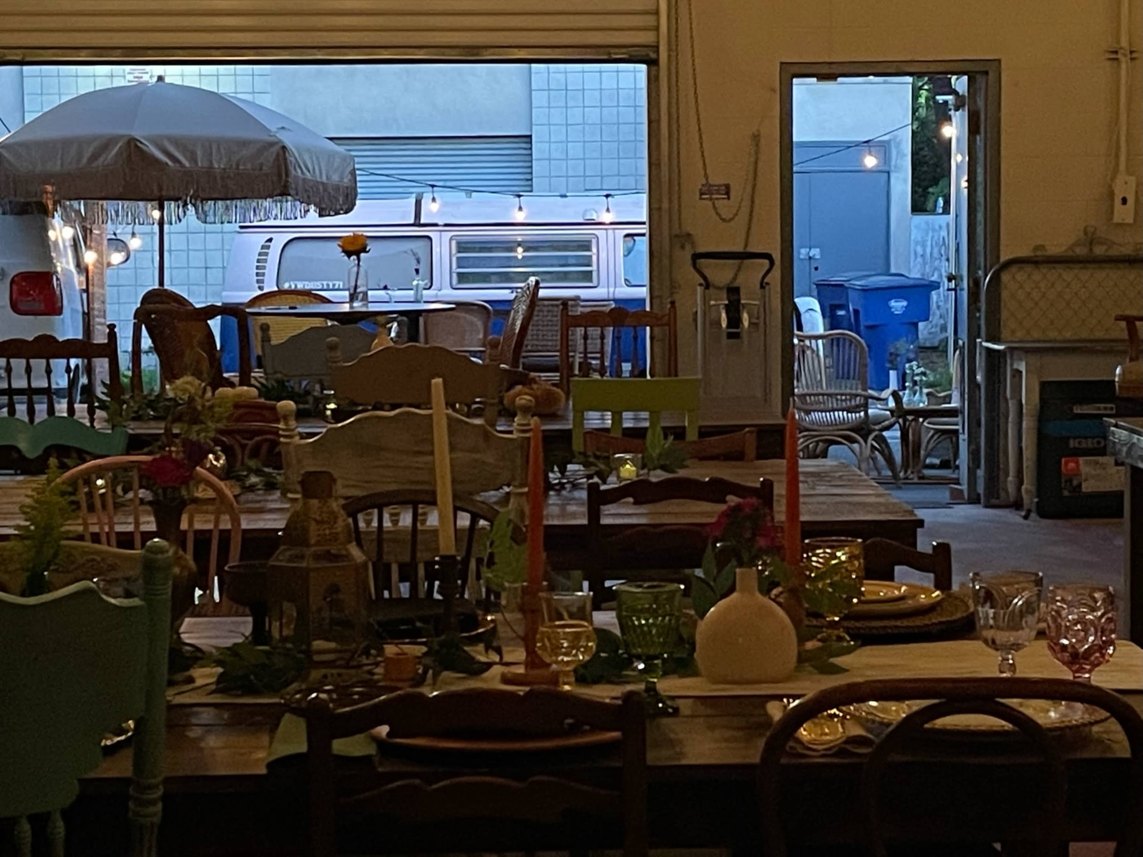 The image shows a collection of tables set with various decorations and tableware, with a vintage van visible through an open doorway in the background.