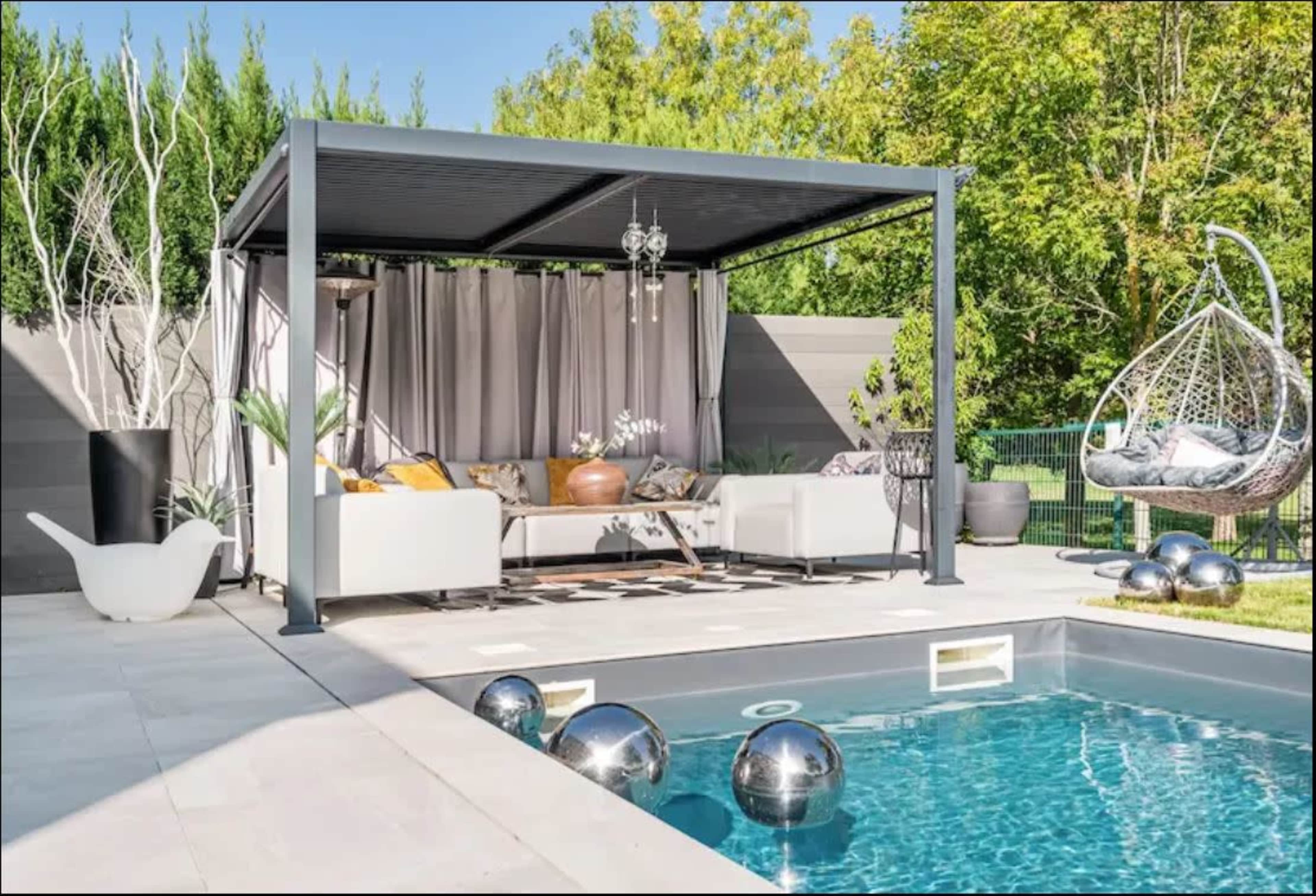 The image shows a modern outdoor lounge area with a covered seating arrangement next to a swimming pool, surrounded by greenery and decorative elements.