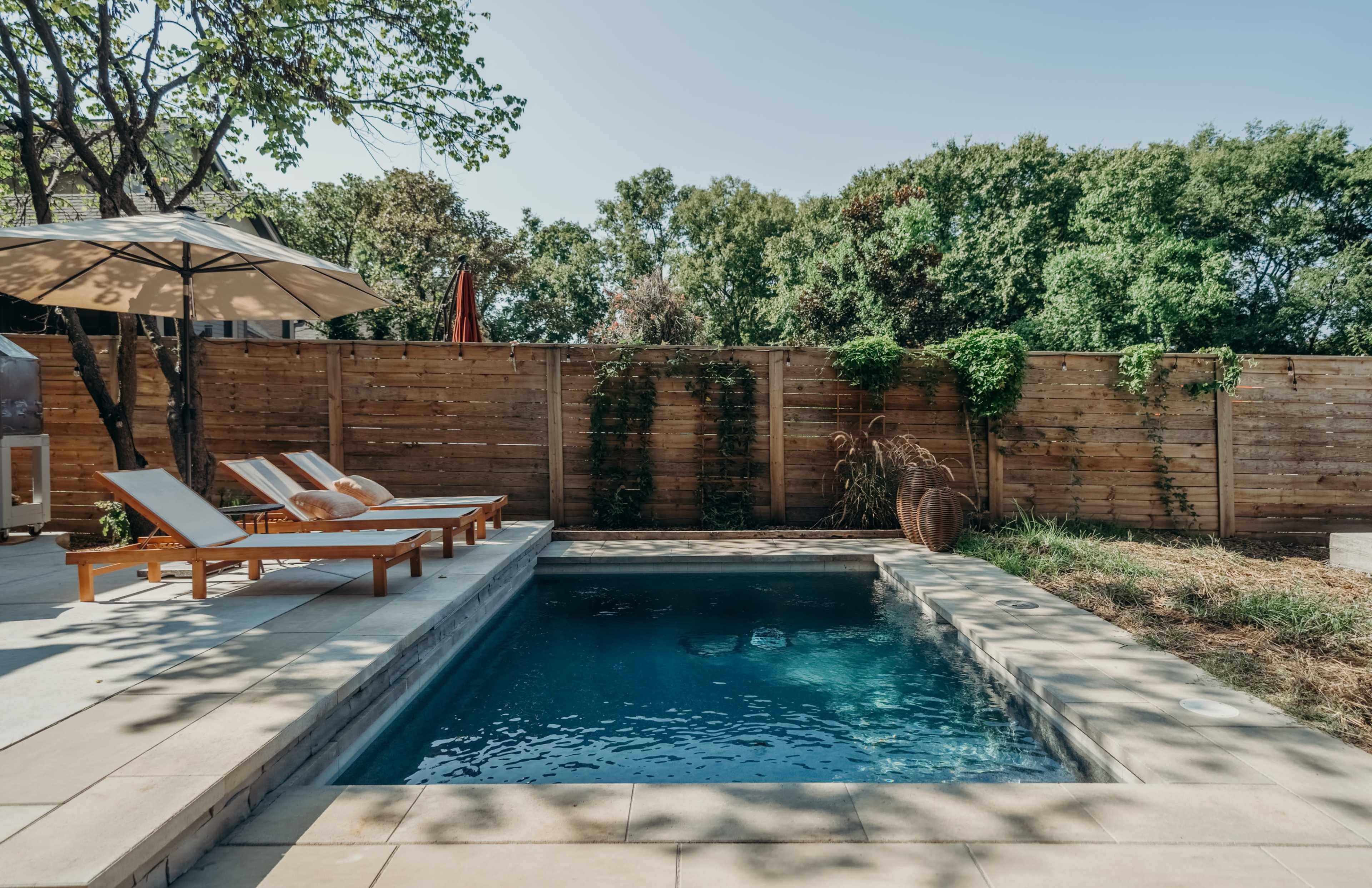 A rectangular swimming pool is surrounded by wooden decking and lounge chairs under a large umbrella, with a wooden fence and greenery in the background.