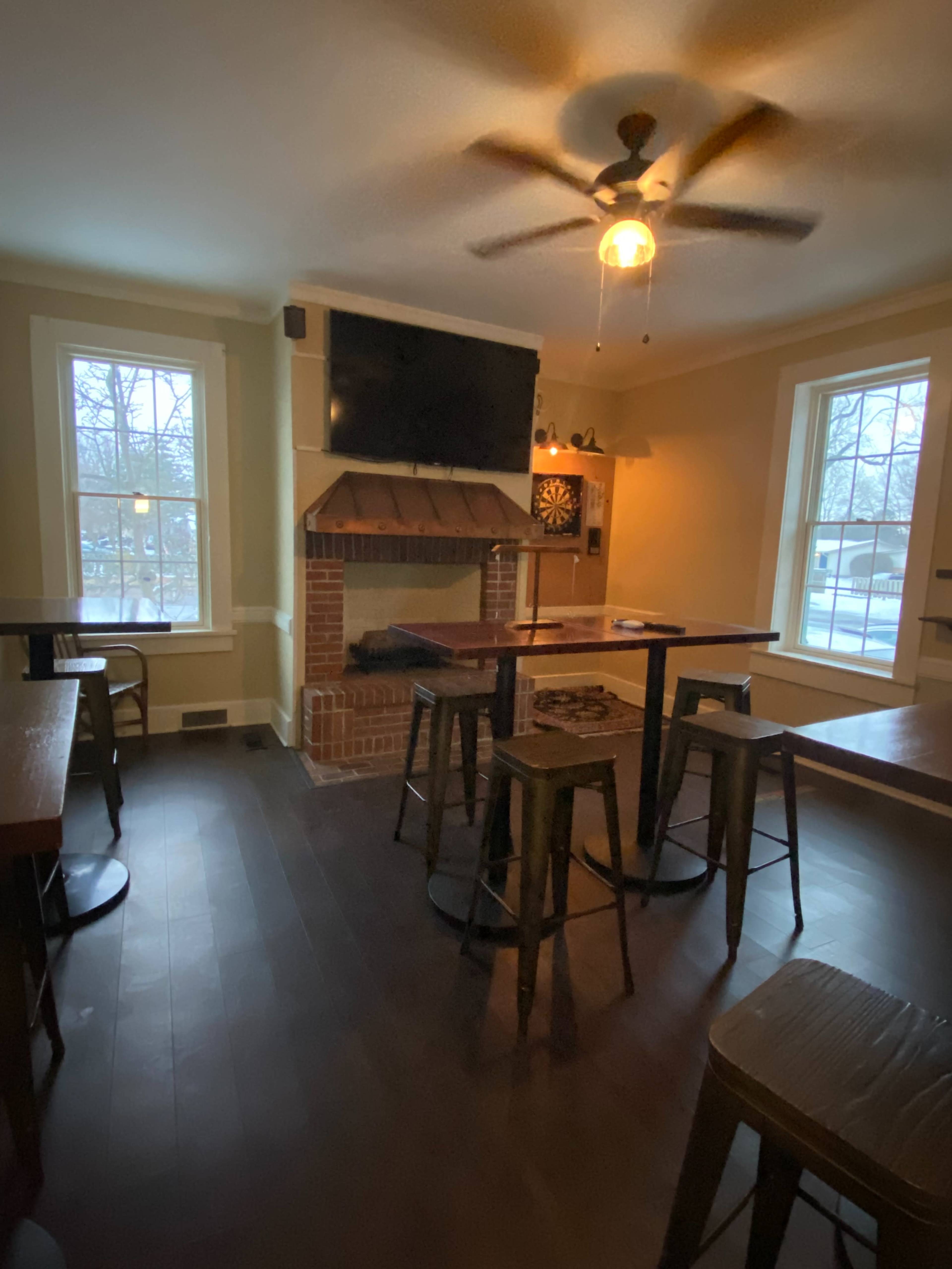 The image shows a cozy room with a fireplace, a ceiling fan, and several tall tables and stools arranged around the space.