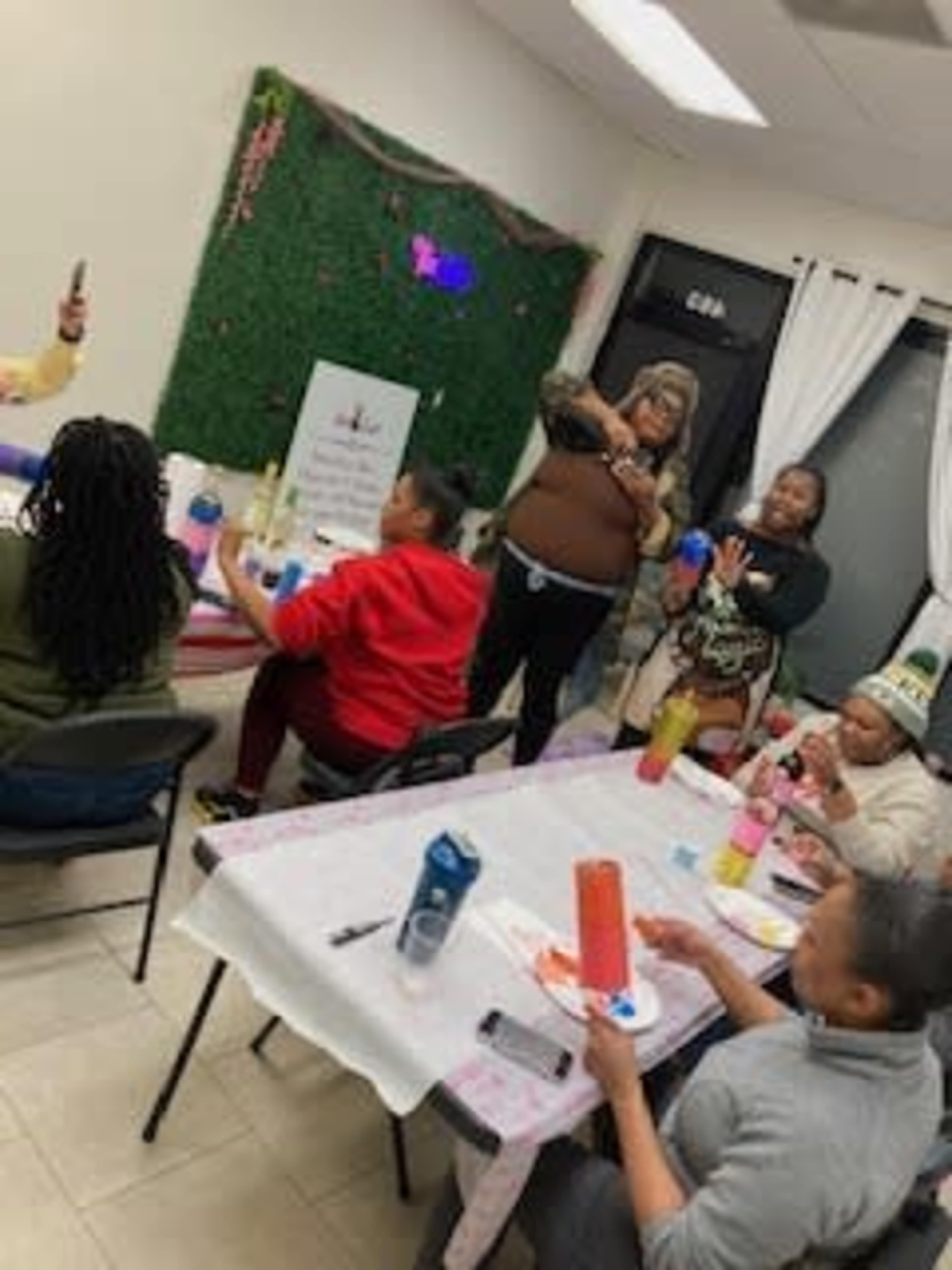 A group of people are engaged in a craft activity around tables in a brightly lit room decorated with a green wall display.