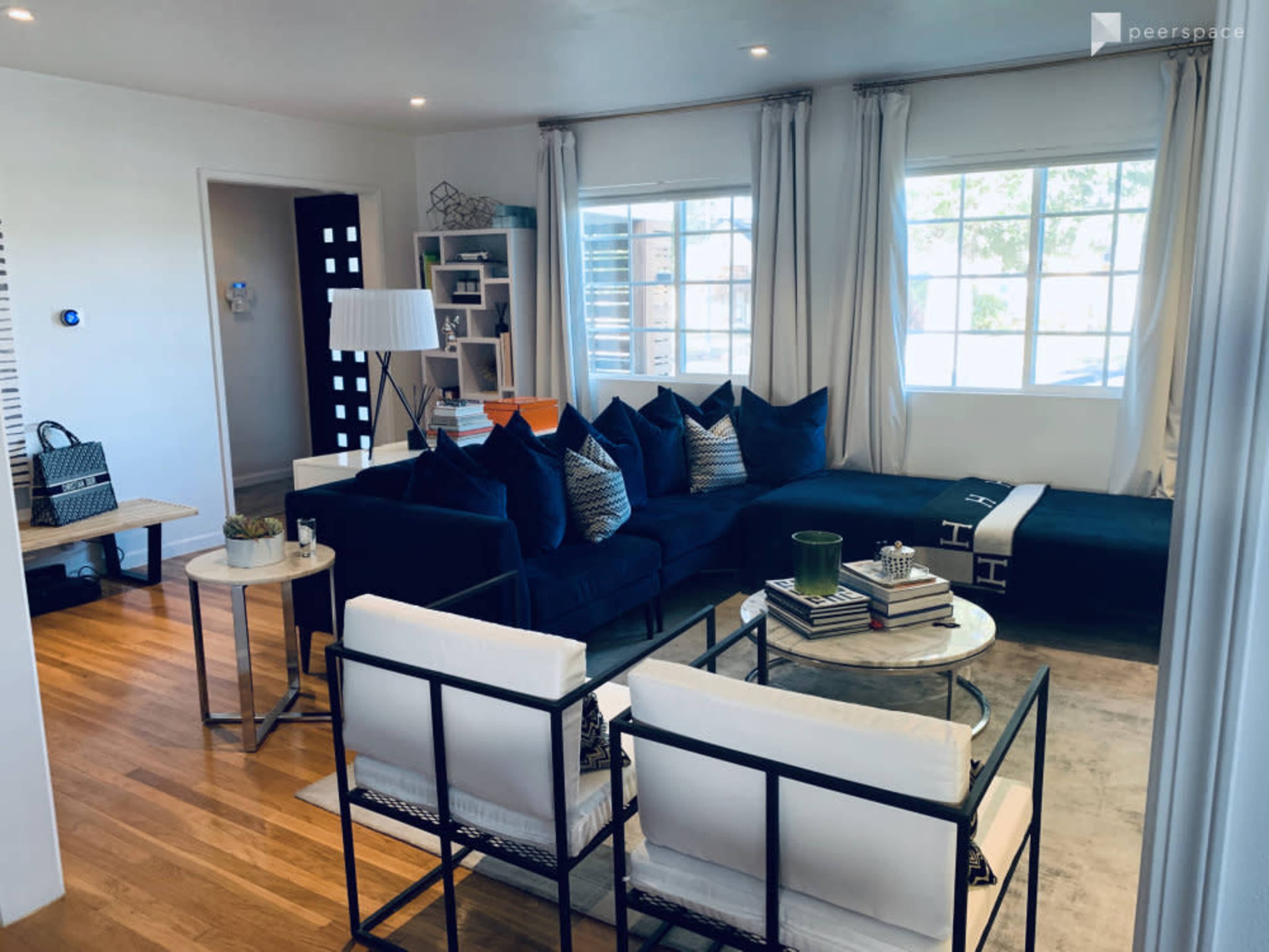 A modern living room featuring a dark blue sectional sofa, two white chairs, a coffee table, and large windows allowing natural light.