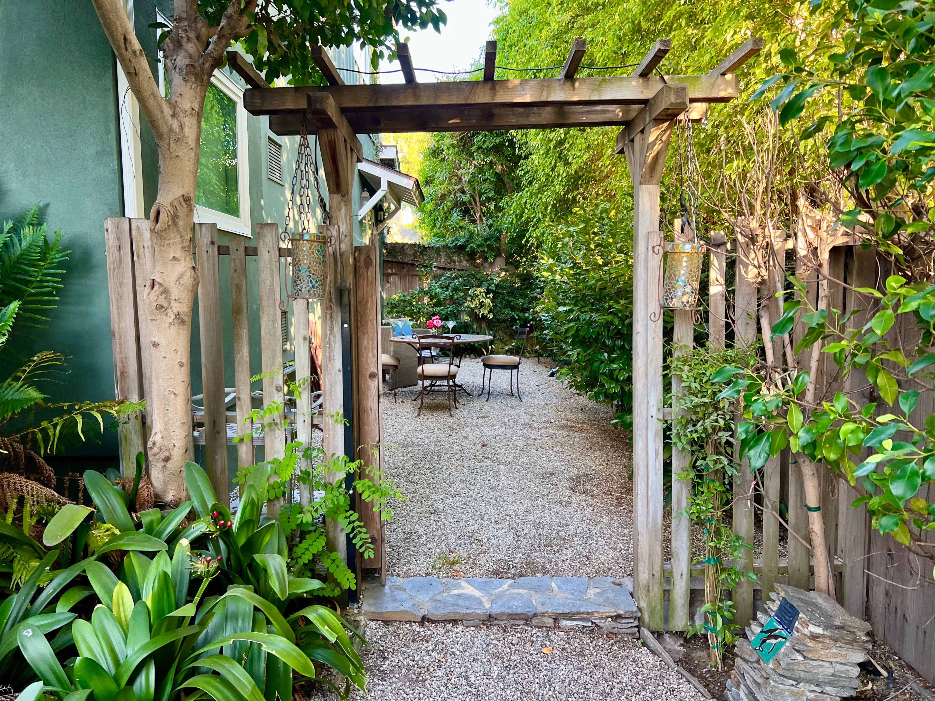 A gravel pathway leads through a wooden archway into a garden space with seating surrounded by greenery.