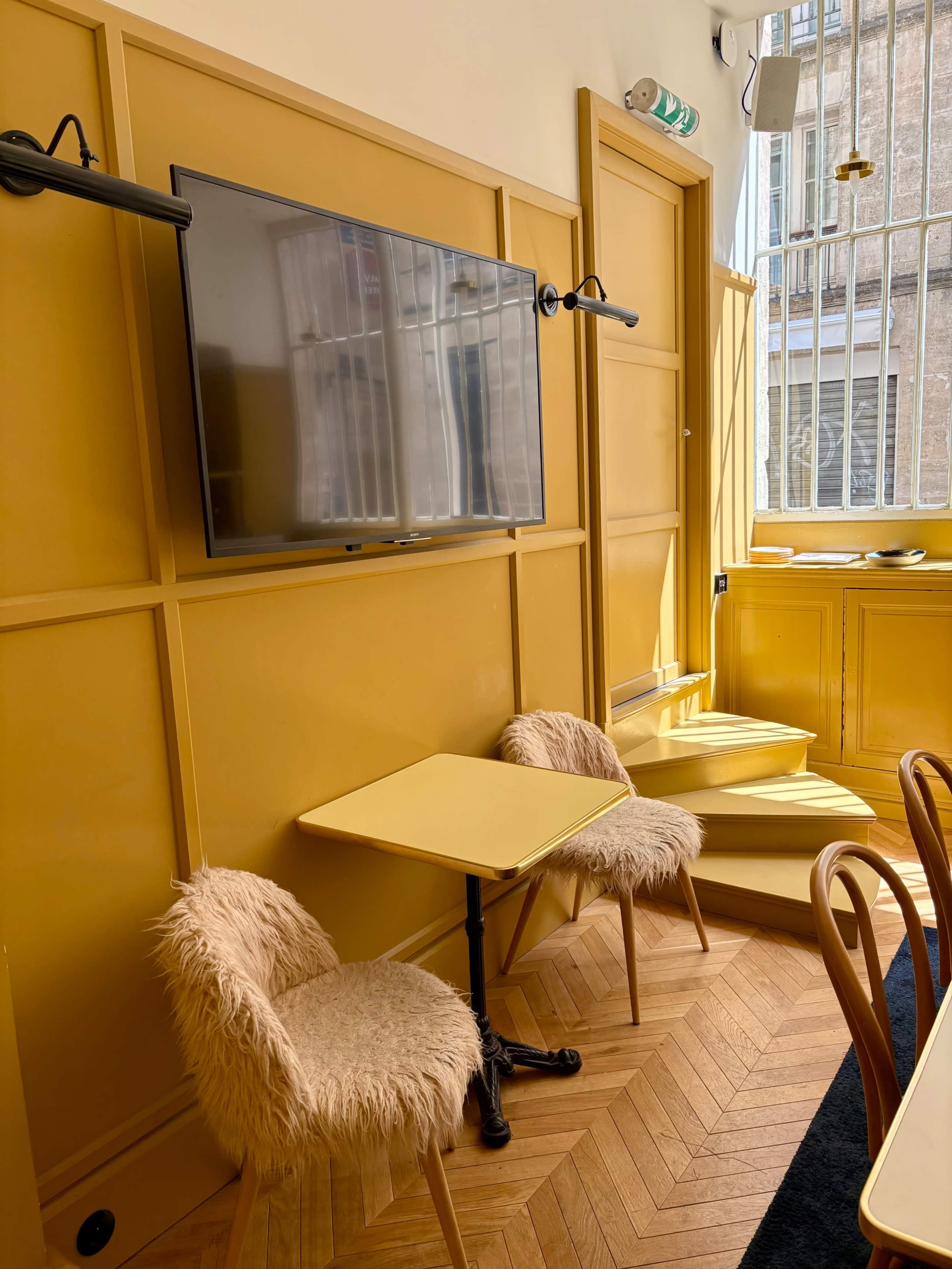 A cozy room features two fluffy chairs and a small table near a wall-mounted television, with yellow walls and a staircase in the background.