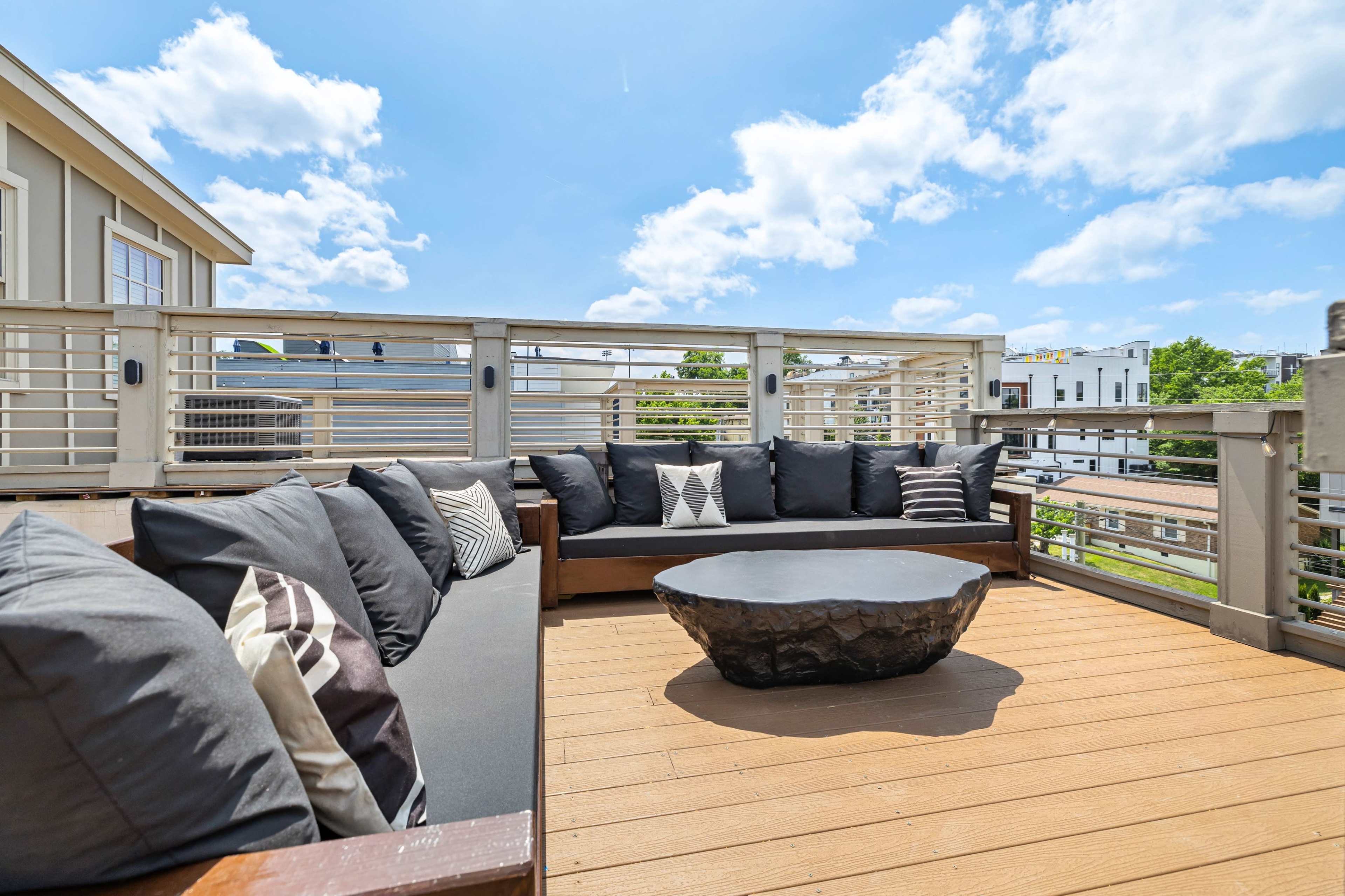 The image shows a rooftop patio with a sectional couch and a stone coffee table, surrounded by a railing and a clear blue sky.