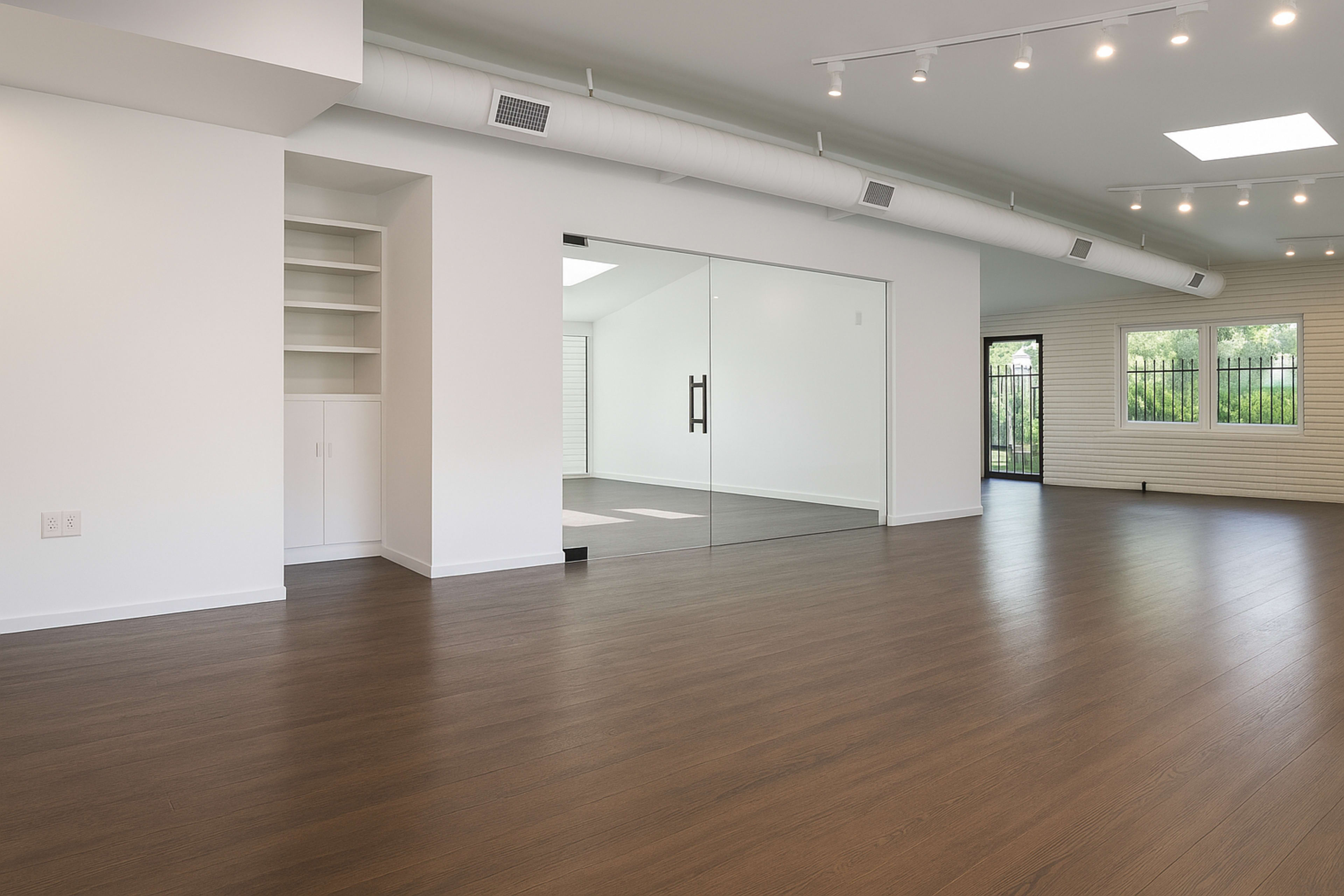 The image shows a spacious, modern interior room with light-colored walls, a large glass door, and wooden flooring.